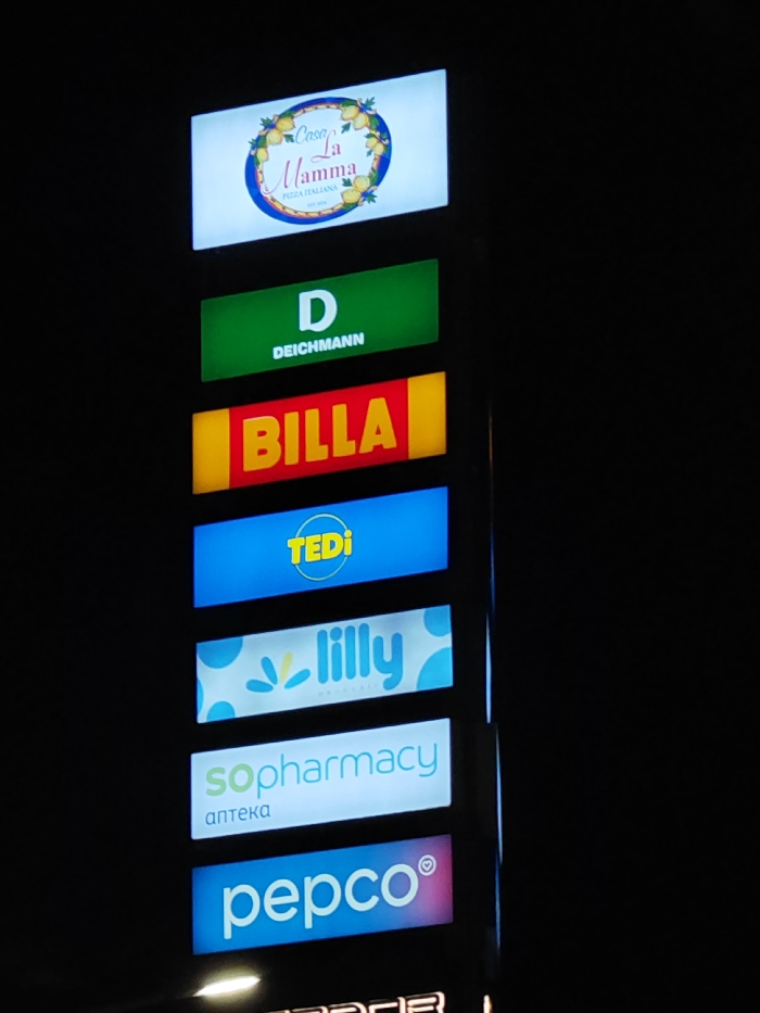 The image displays a vertical, illuminated pylon sign against a dark, nighttime background. The sign is composed of seven distinct, rectangular backlit panels stacked vertically, each presenting a brand logo. From top to bottom, these logos are:
1. **Casa La Mamma:** A white panel featuring a logo with "Casa La Mamma" in blue script, "PIZZA ITALIANA" in smaller blue text, all framed by a wreath of yellow lemons on a blue circular border.
2. **Deichmann:** A green panel with a large white "D" above "DEICHMANN" in smaller white text.
3. **BILLA:** A yellow panel displaying "BILLA" in prominent red block letters.
4. **TEDI:** A blue panel featuring "TEDI" in yellow text, enclosed within a yellow oval outline.
5. **lilly drogerie:** A white panel with "lilly" in stylized light blue font, accompanied by light blue droplet-like graphical elements and "drogerie" in smaller text below.
6. **sopharmacy:** A white panel showing "sopharmacy" in light green text, with the Cyrillic word "аптека" (apteka) in smaller grey text below.
7. **pepco:** A blue-to-purple gradient panel featuring "pepco" in white lowercase letters, followed by a small heart symbol.
The pylon sign is located at Grand Plaza Shumen, Tsar Osvoboditel Blvd, Shumen, Bulgaria.