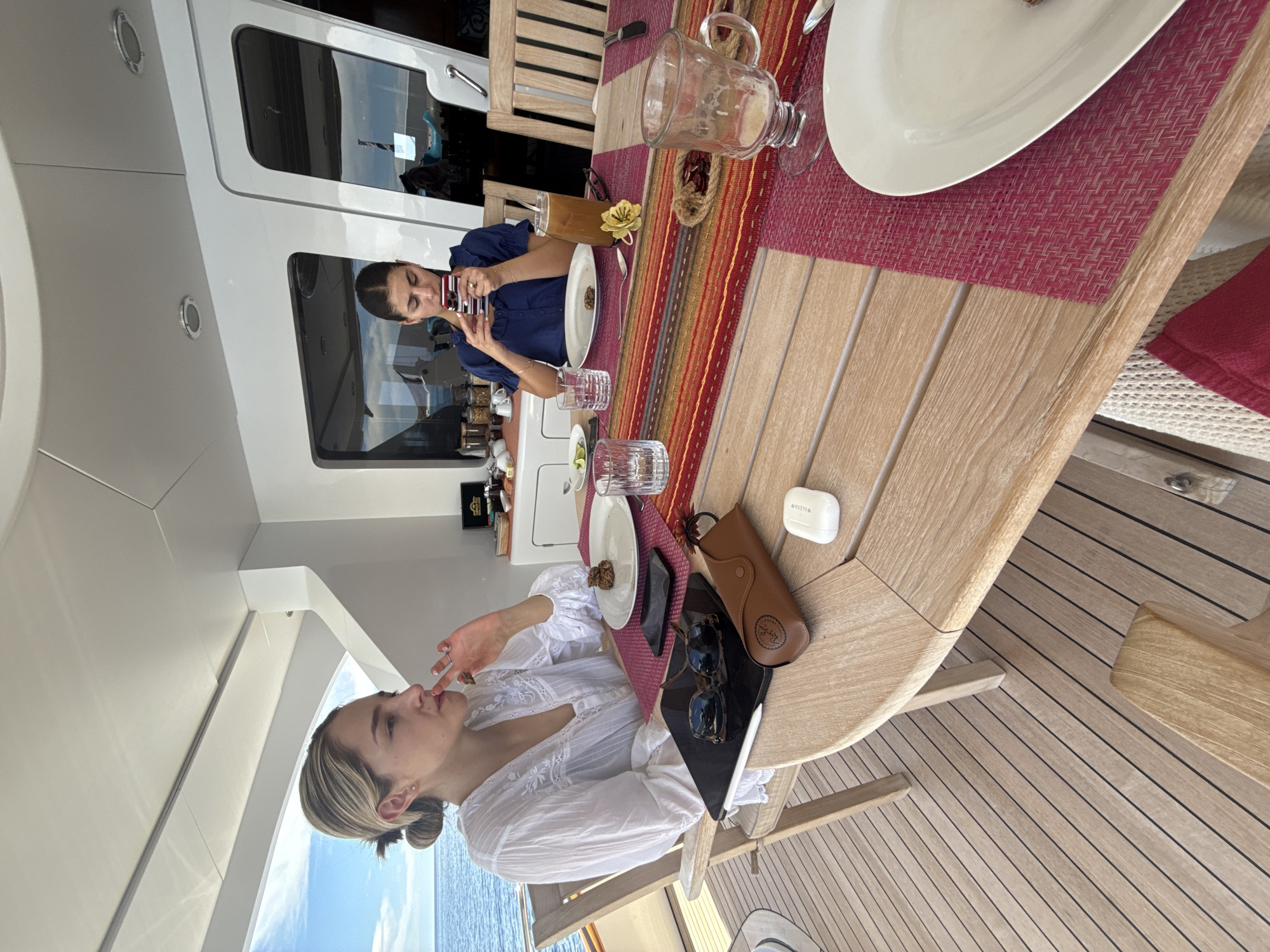 Two women are at a table on a boat. One woman, wearing a white blouse, is sitting at the table eating. She has a tablet next to her with a pair of sunglasses and a glasses case on top of it. The second woman, wearing a blue shirt, is taking a picture with her phone. The table has a red, orange, and yellow placemat, and a variety of plates and glasses. The boat is sailing on the ocean near Unknown, Mexico.