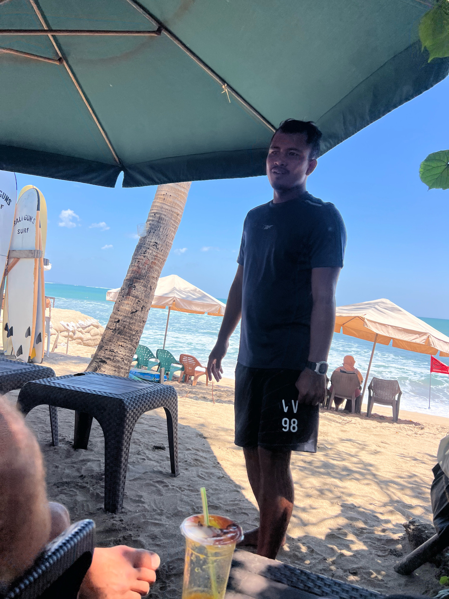 The image depicts a beach scene in Kuta, Indonesia, on a sunny day. A man stands beneath a large green umbrella, wearing a dark t-shirt and black shorts with white lettering that reads "VV 98". He appears to be looking towards the right of the frame.
In the background, the ocean is visible with gentle waves, and the sky is a clear blue. Several beach umbrellas are set up on the sand, with chairs beneath them. Two surfboards are propped up near a palm tree, one of which has "BALI GUN.S SURF" written on it. In the foreground, a plastic table with a woven texture sits on the sand, and a person's foot is visible resting on a chair. A plastic cup with a straw and a beverage is placed on the table, catching the sunlight. The overall scene suggests a relaxed atmosphere at a beachside establishment in Kuta.