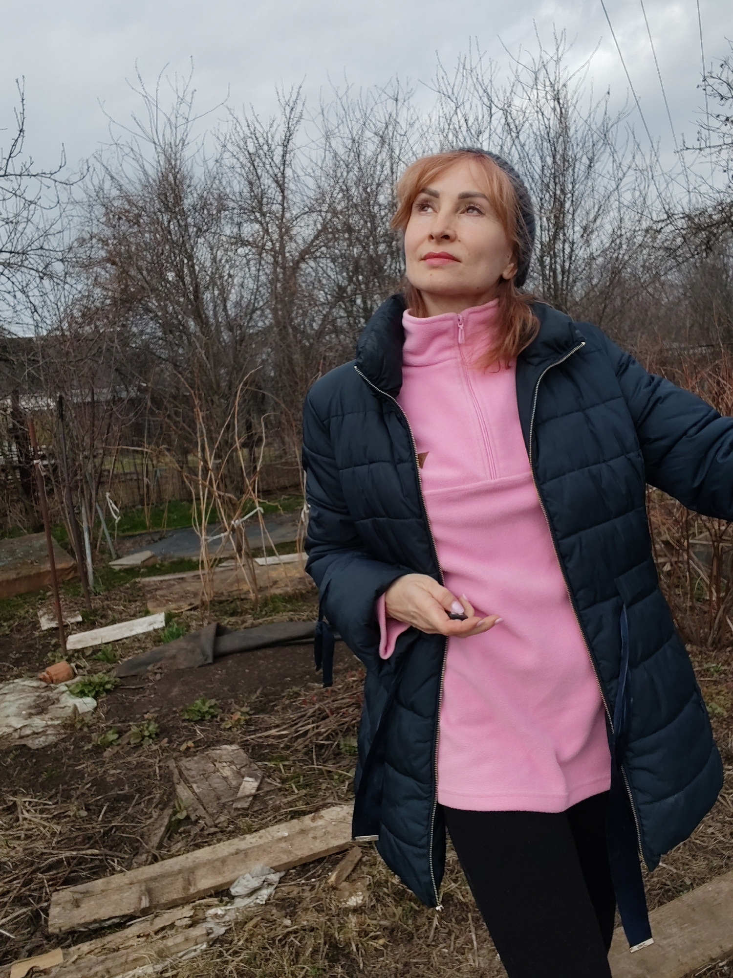 A woman stands in a cluttered garden, likely in the city of Chaikovsky, Russia, given the location metadata. She wears a dark blue quilted jacket over a pink fleece with a partial zipper. She also wears black leggings. A gray beanie covers some of her reddish-blonde hair. She is looking upward with a thoughtful expression. The background consists of numerous leafless bushes and trees against an overcast sky. The ground is covered in dead grass, weeds, and scattered pieces of wood and debris. A fence is visible in the background, further delineating the garden space. The woman holds a small dark object in her hand.