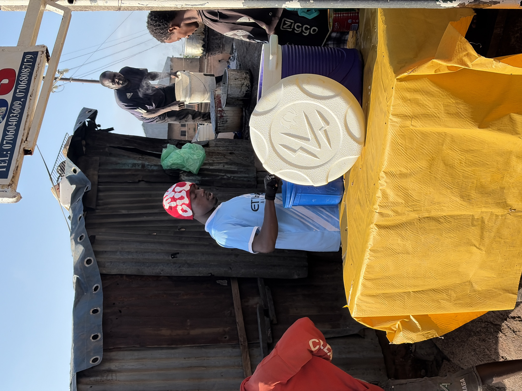 In Gura Top, Nigeria, under a bright sky, a food stall is set up. The stall has a corrugated metal structure with a tarp over the top. A yellow cloth covers a table in front. A young man in a blue shirt and a red and white hat stands behind the table, looking down at his hands which are obscured by a blue container. To his right, a young woman in a dark shirt with a zig-zag pattern watches him. In the background, a man in a dark shirt is holding a bucket, with steam rising around him. There are several other containers and cooking pots around the stall. The overall scene suggests a busy, open-air food preparation and selling environment.