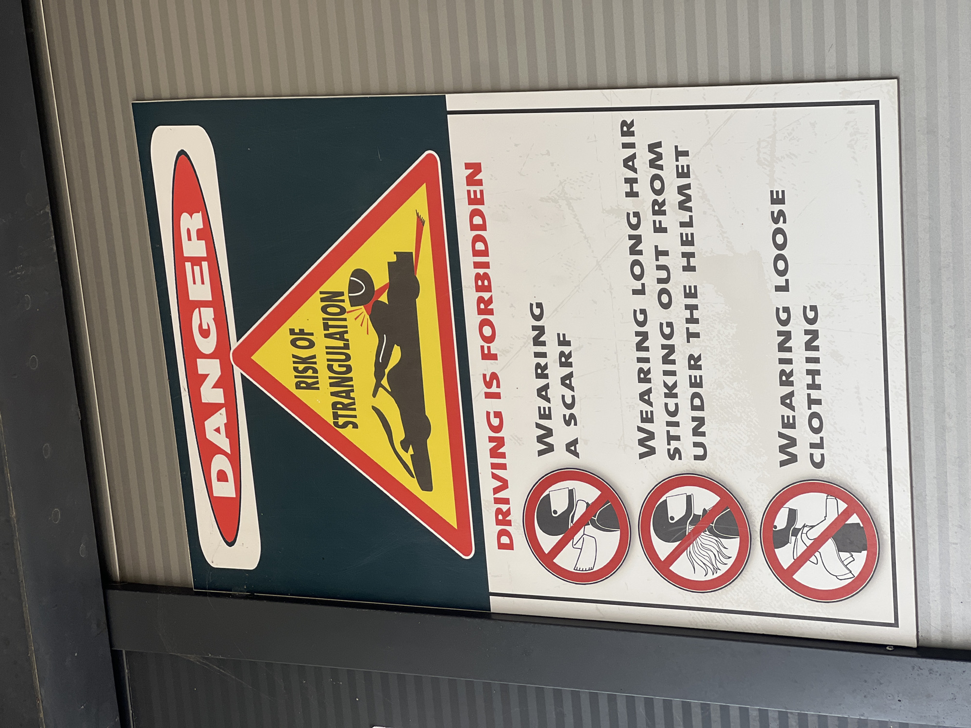 The image shows a safety sign that reads "DANGER RISK OF STRANGULATION DRIVING IS FORBIDDEN". It lists and shows illustrations of what is forbidden while driving, including wearing a scarf, wearing long hair sticking out from under the helmet, and wearing loose clothing. The location in the city of Bigor, Montenegro is unknown.