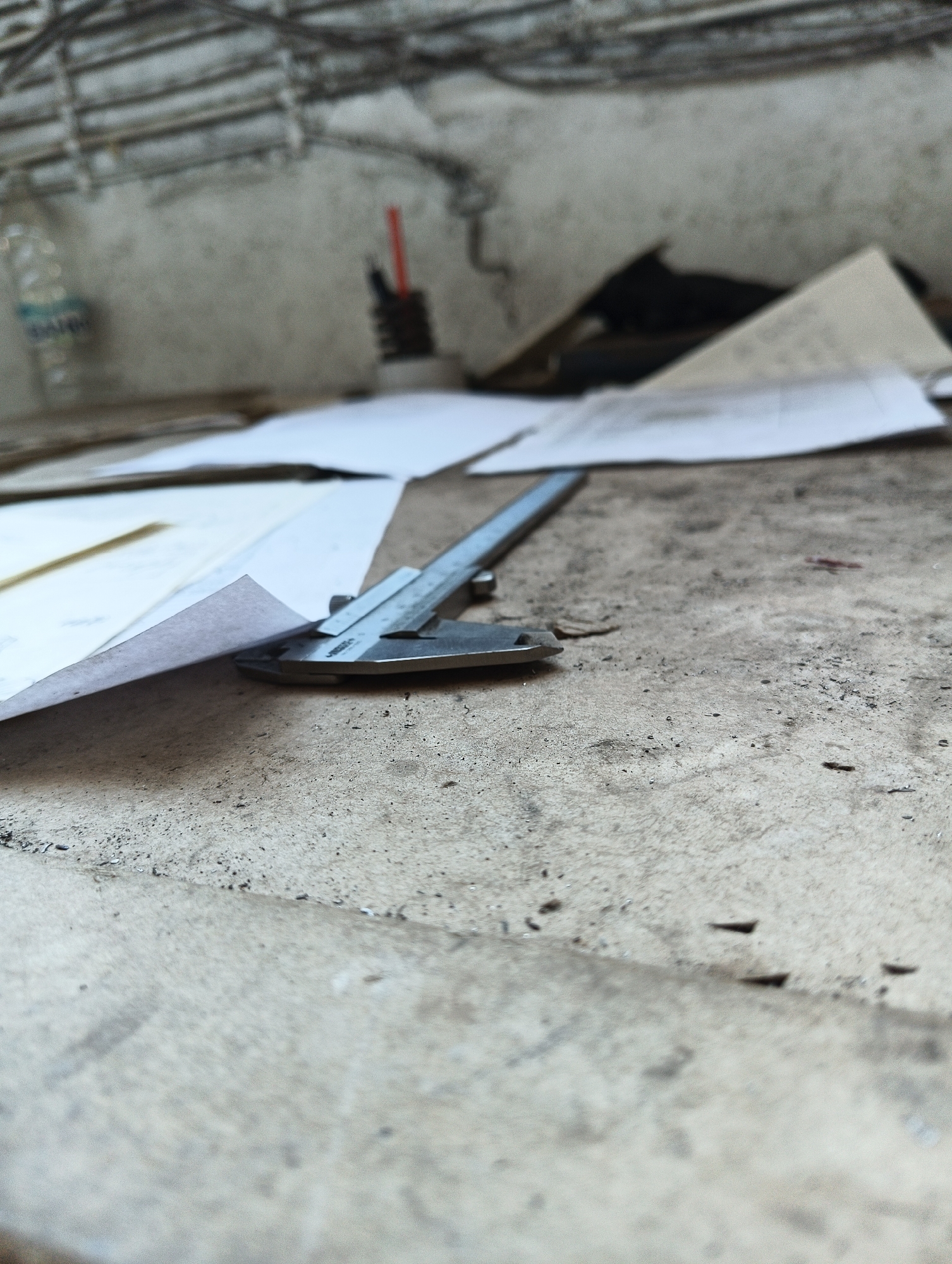 The image shows a close-up, low-angle shot of a metal caliper resting on a textured, dusty concrete surface. Several sheets of paper are spread around the caliper, with one sheet folded and partially covering the tool. The background is out of focus but appears to be a rough concrete wall with some electrical conduits or pipes running across the top. A few small dark objects are scattered on the concrete in the foreground and mid-ground. A blurry plastic bottle is visible in the upper left corner. The scene suggests a workspace or construction area. The location in Karnobat, Bulgaria is not visually discernible from the image itself.