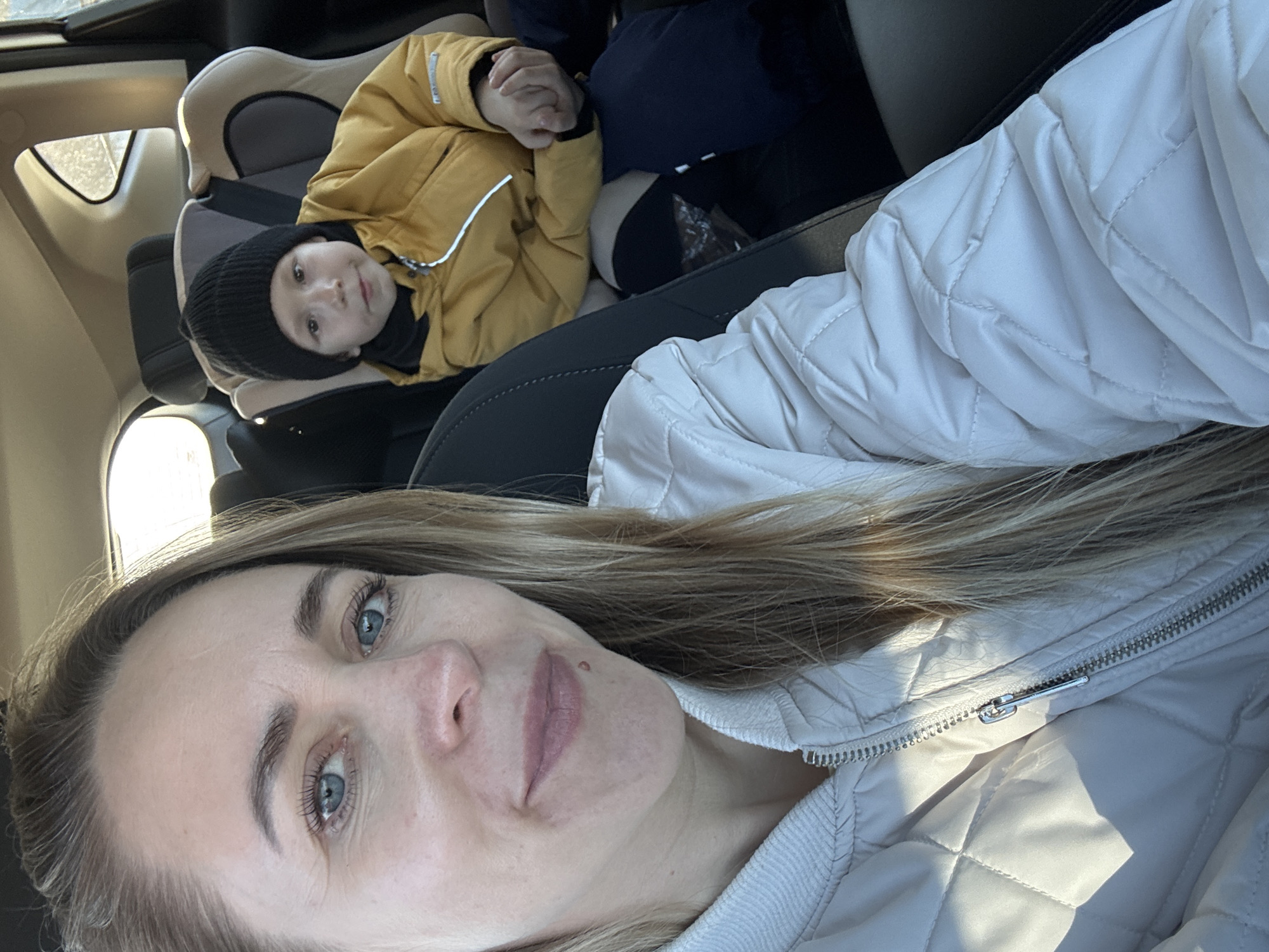 The image shows a woman taking a selfie in a car. A young child in a black beanie and yellow jacket is seated in a car seat behind her. The location is likely Chaikovsky, Russia, but there is no visual confirmation in the image to definitively confirm the location. The woman is wearing a quilted white jacket.