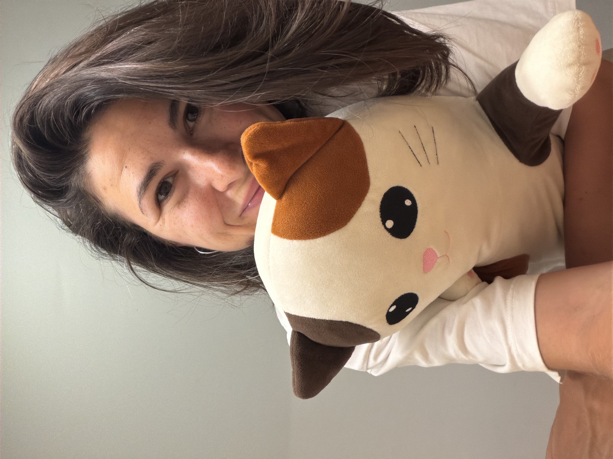 A woman with shoulder-length brown hair and fair skin is holding a stuffed cat. The woman is wearing a white shirt and has an earring on her left ear. The stuffed cat is cream-colored with brown patches and black eyes. The background is a plain gray wall. There is no indication of Budva, Montenegro, or any specific location.
