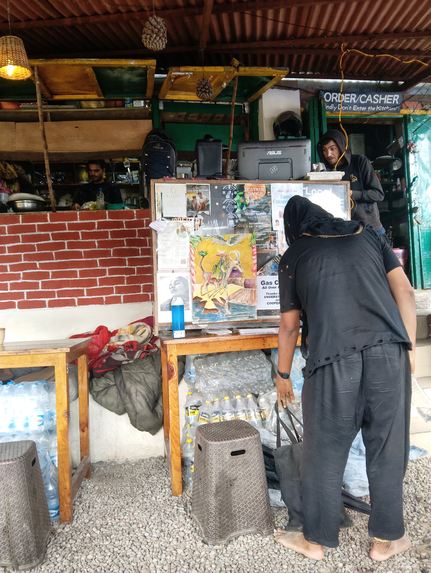 The image depicts an outdoor scene at a small shop, likely in Haini, India. In the foreground, a person wearing a black headscarf and a black, tattered t-shirt and pants stands facing away from the camera, looking at a display board. Their left hand is resting on a black fabric bag. To the left and right of this person are brown, woven stools. Behind them, wooden tables are visible, stacked with numerous plastic bottles of water and other beverages. A gray blanket and a green garment are also seen on the tables.
The display board is mounted on wooden supports and features various posters and papers, including what appears to be a flyer for "THAI YOGA MASSAGE" and another with text about "Gas". A blue aerosol can is placed on a lower shelf of the display.
In the background, a man in a black hoodie is visible behind a counter, interacting with what appears to be a cash register or payment terminal. A printer is also on the counter. Above the counter, shelves are stocked with goods, and a man in a blue shirt is visible working in the back. The shop has a corrugated metal roof supported by wooden beams. The ground is covered with small gray pebbles. A sign above the counter reads "ORDER/CASHER Kindly Don't Enter the Kitchen".