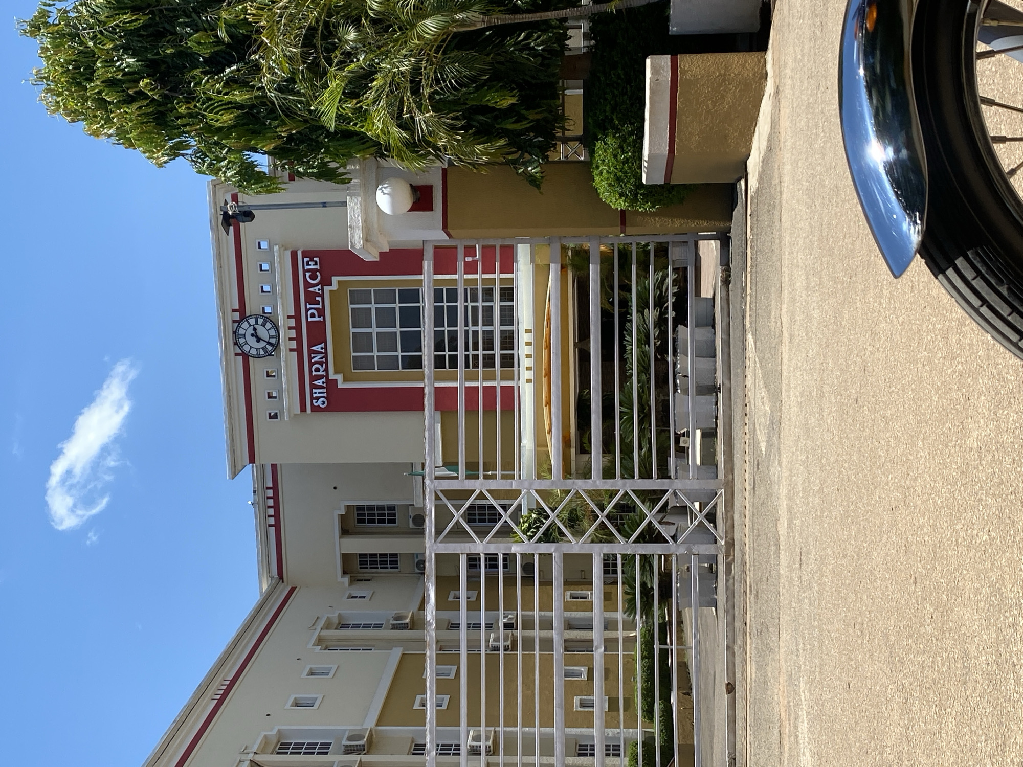 The image shows the entrance to "SHARNA PLACE" in Jos, Nigeria, on a sunny day. A large building with yellow and red accents is visible behind a white metal gate. The building has multiple windows and air conditioning units. A clock is mounted on the facade above the building's name. To the right of the building, there is a tall green tree and a palm tree. In the foreground, part of a motorcycle's chrome fender and wheel are visible, suggesting the image was taken from a motorcycle. The ground in front of the gate is paved with asphalt.