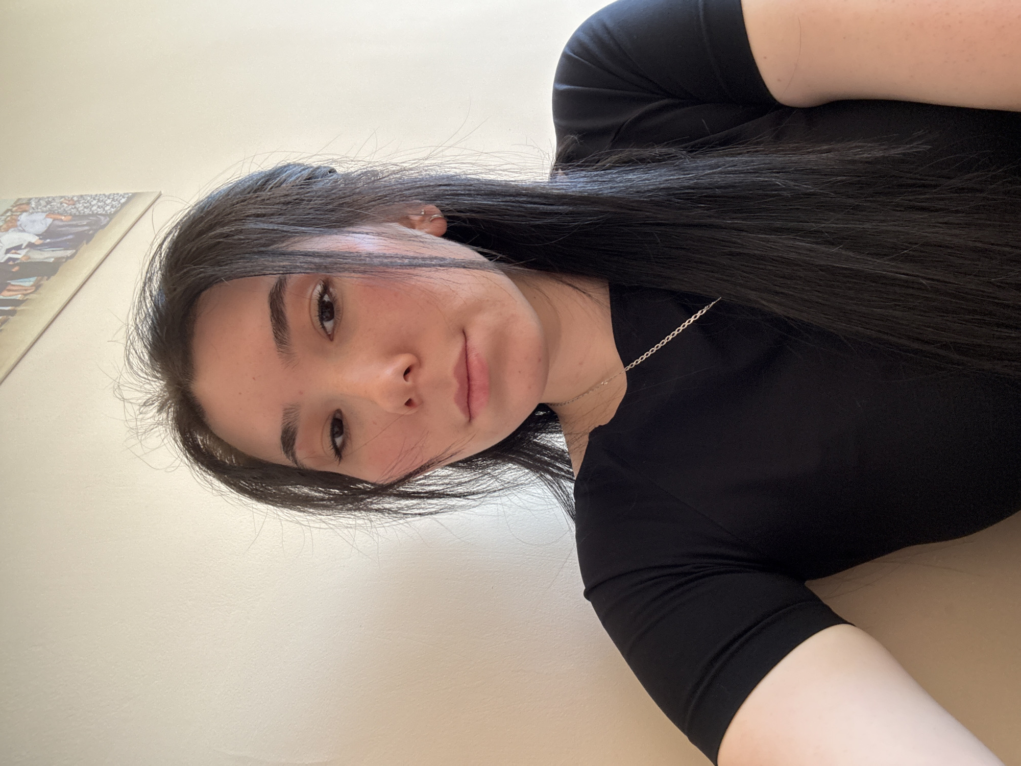 A woman with dark hair is looking directly at the camera. She has fair skin, dark eyebrows, and a small silver necklace. She is wearing a black shirt. The background is a pale yellow wall with a framed picture on the left. This image was not necessarily taken in Izhevsk, Russia.