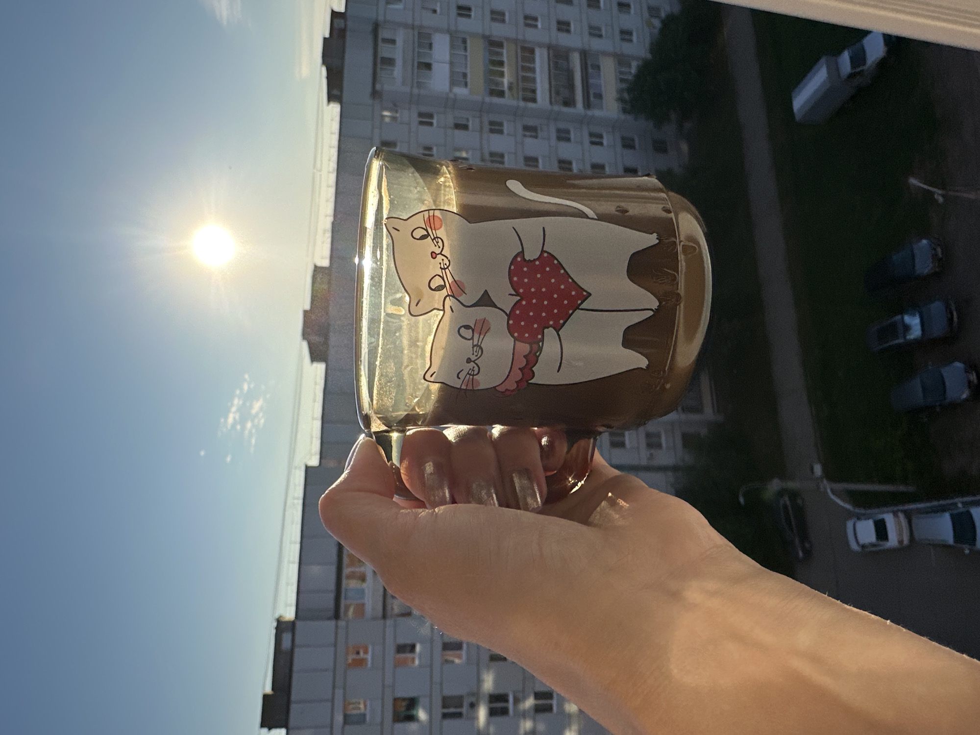 A person's hand, adorned with metallic silver nail polish, holds a clear glass mug filled with a dark liquid, likely coffee or tea. The mug features a cartoon illustration of two white cats embracing, one holding a red heart with white polka dots. The mug is held up against a bright sun with visible rays, casting a glare on the glass. In the background, a multi-story apartment building with numerous windows is visible, suggesting an urban setting. Below the building, a road and parked cars can be seen. The overall scene captures a moment of enjoying a drink outdoors, likely on a balcony, with a clear blue sky and the sun prominent. The location is identified as Chaikovsky, Russia.
