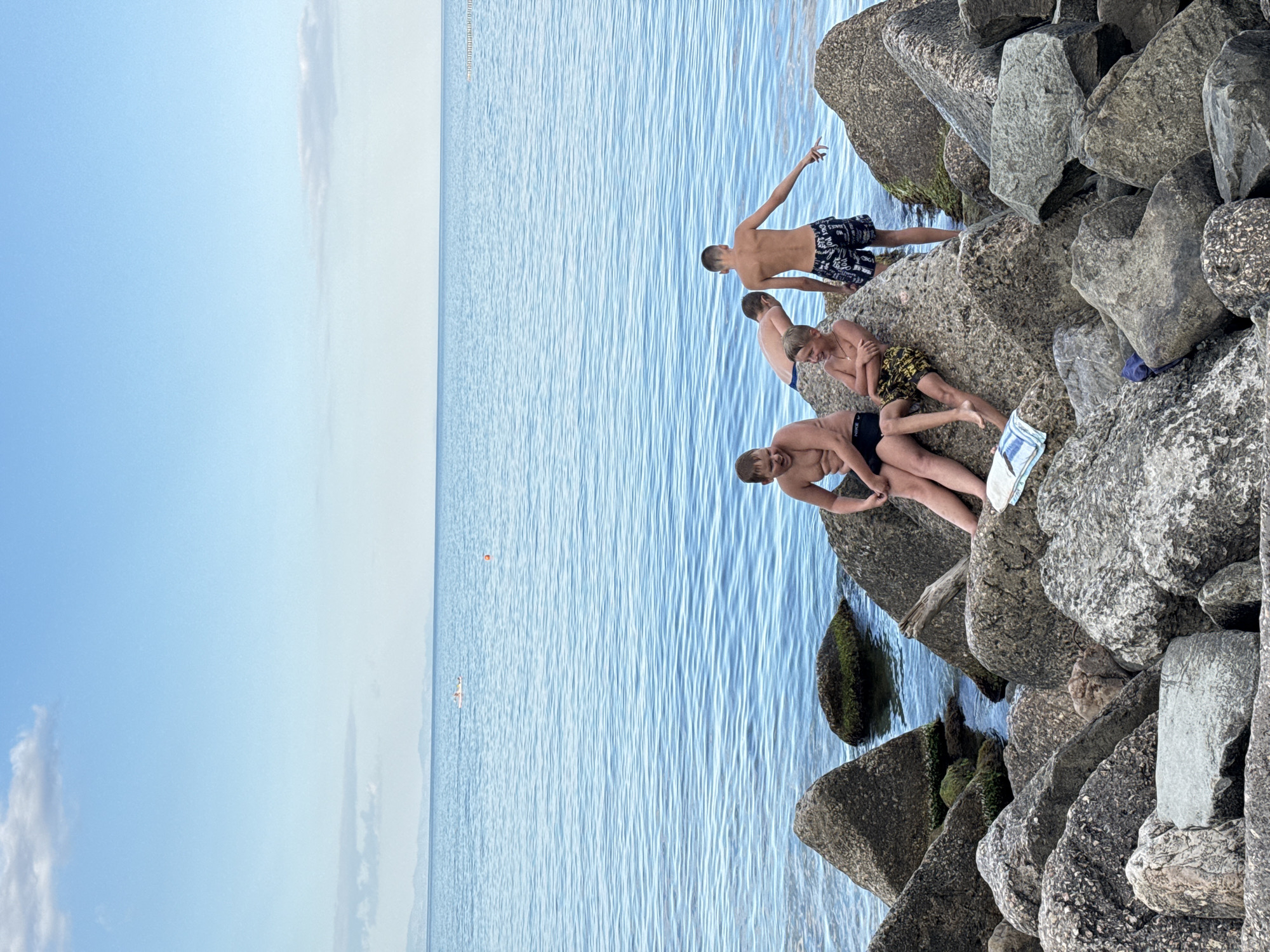Four boys are posing on rocks by the sea in Partenit, Ukraine. Three of the boys are sitting on large rocks, with one boy standing to the right of them, facing away from the camera. All boys are wearing swim trunks. The sea is calm with gentle waves, and the sky is clear and blue with a few clouds. The rocks are large and gray, some covered with moss. The scene appears to be a sunny day at the beach.