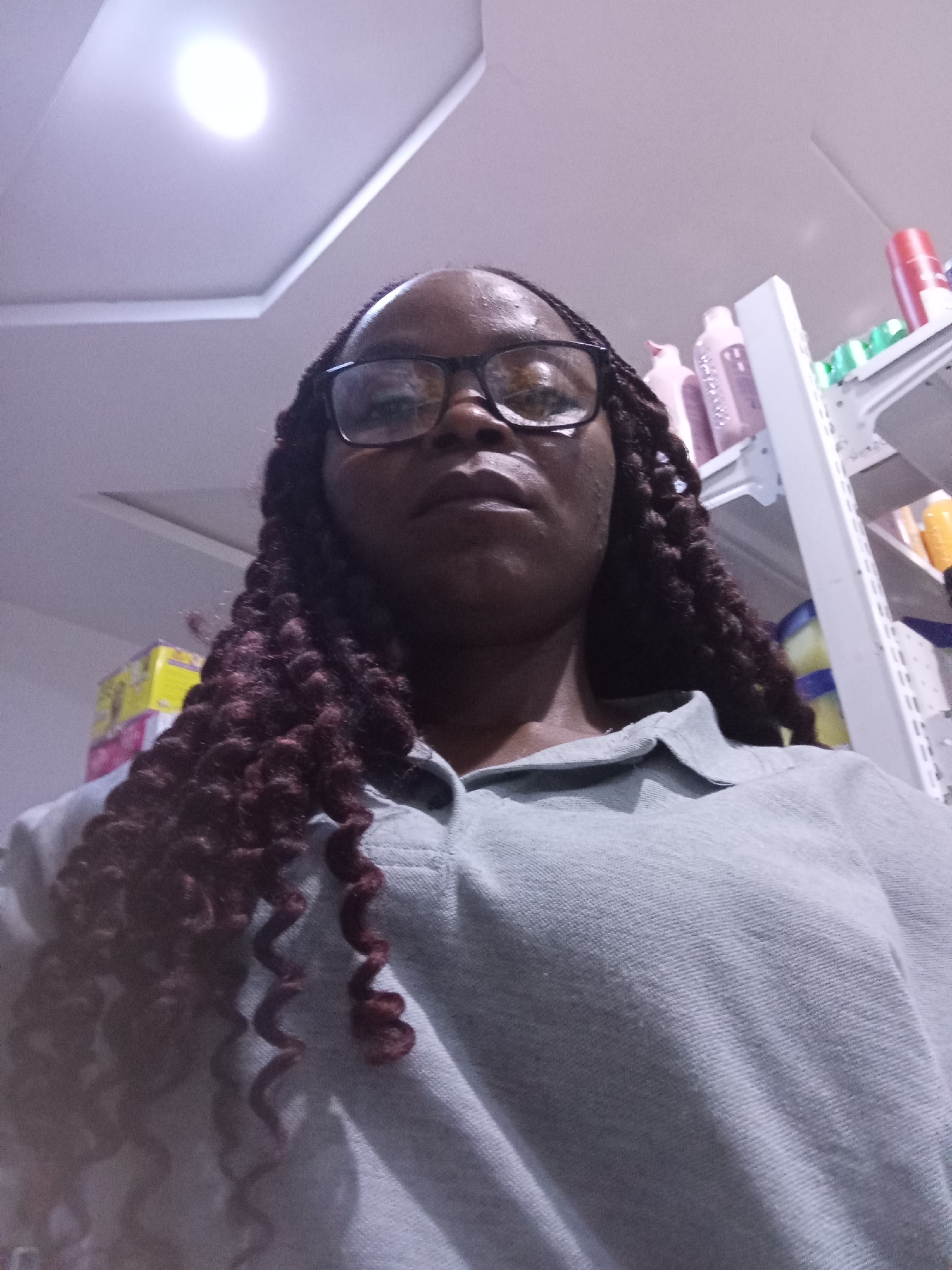 The media file is a close-up, low-angle selfie of a Black woman wearing glasses and a light blue collared shirt. She has dark skin and her hair is styled in long, dark, curly braids that fall over her shoulders and chest. The background appears to be an indoor setting, possibly a store or pharmacy, with shelves stocked with various products.
Notable details include:
- **Setting:** The presence of shelves filled with products, including bottles and boxes, suggests a retail environment. The lighting in the upper left corner indicates an overhead light fixture, possibly in a ceiling.
- **Objects:** Several bottles, some appearing to be personal care or cleaning products, are visible on the shelves in the background. One red bottle stands out on a higher shelf. A yellow and white box is also visible.
- **Time of Day/Weather:** No information about the time of day or weather can be inferred from the image.
- **Emotions:** The woman's expression is neutral, and she is looking directly at the camera.
There is no visible text in the image. The overall impression is a casual, personal photograph taken in a retail setting.