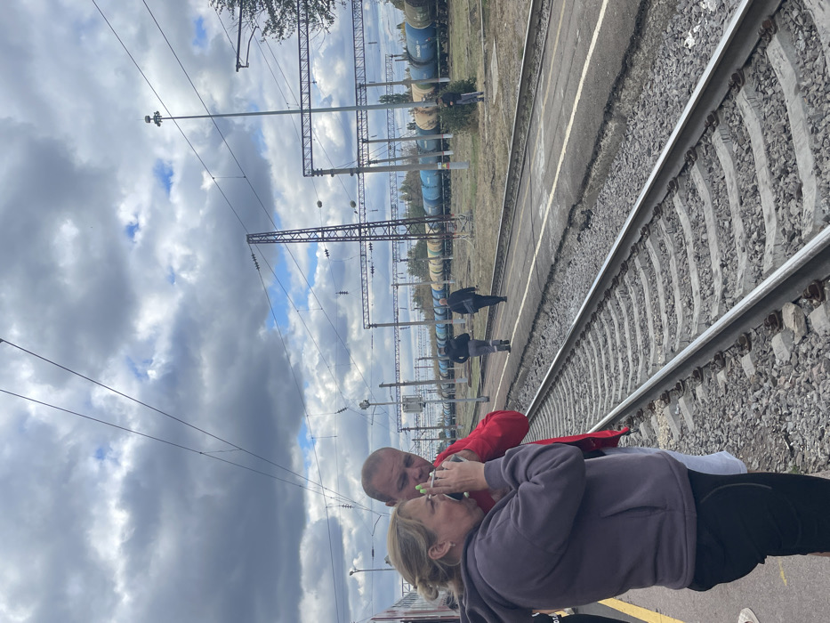The image portrays a railway environment in Gryazi, Russia, under a sky with prominent gray clouds and patches of blue. In the immediate foreground, on the left, a woman with blonde hair, styled back, is seen wearing a dark gray hooded sweatshirt and dark pants. She holds a bottle with a green cap to her mouth, appearing to drink from it. Her fingernails are painted light green. Standing slightly behind and to her right is a man with a bald or closely shaven head, dressed in a red long-sleeved shirt and white shorts. Both are positioned on a train platform with a yellow safety line visible on the far left. Adjacent to the platform are railway tracks featuring concrete sleepers and gravel ballast.
In the midground, to the right of the foreground tracks, is another platform or walking path. Two male figures are observed walking away from the camera along this path; one wears a dark jacket over light pants, and the other is in dark attire. Further in the background and to the right, a third male figure stands near multiple sets of railway tracks. A long line of stationary freight cars, consisting of blue and yellow tanker wagons, extends across the background. The railway infrastructure includes numerous overhead catenary lines, tall support poles, and lattice towers. A prominent tall light pole with a device mounted on top is visible towards the right midground. Trees and sparse vegetation are discernible beyond the tracks in the distance.