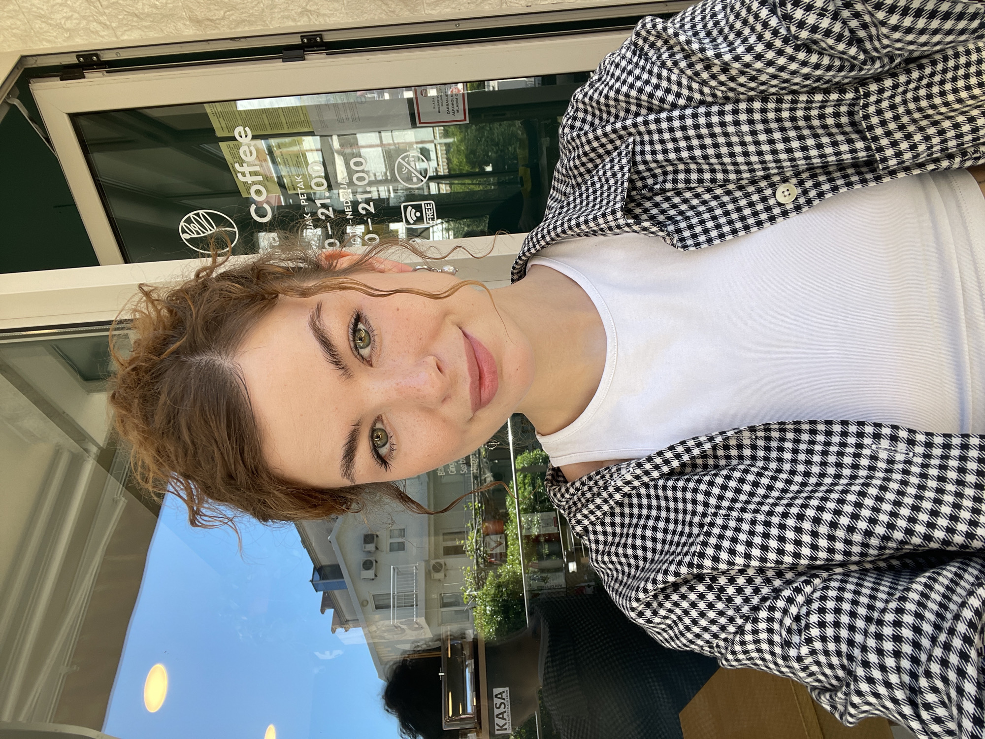 A young woman with curly brown hair and freckles, wearing a white tank top and a black and white checkered button-down shirt, is taking a selfie in front of a coffee shop. The reflection in the glass shows a building with balconies, indicating an urban setting, and the word "Coffee" is visible on the shop's window. The sign indicates that it's located in Budva, Montenegro. The sign also shows the shop's business hours for weekdays and Sundays, and a "Free Wifi" sticker is visible.