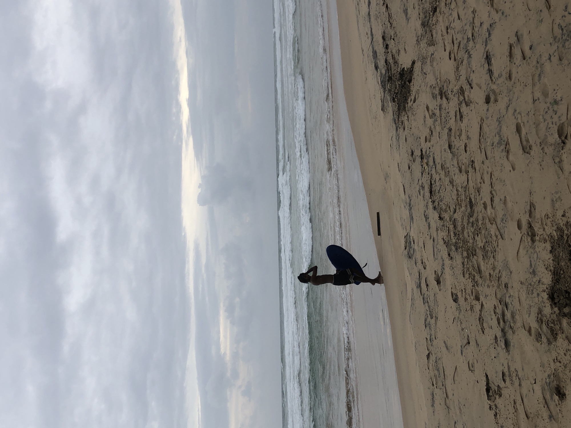 A lone figure stands on a sandy beach in Kathaluwa, Sri Lanka, holding a blue surfboard. The person is silhouetted against the ocean, with their hand raised to their head. The water is choppy, with waves rolling in towards the shore. The sky is overcast, with clouds covering the entire frame. The beach is wet and sandy, with footprints visible in the foreground. There is a piece of driftwood lying on the sand near the figure. The scene appears to be set in the late afternoon or early morning, given the low light conditions.