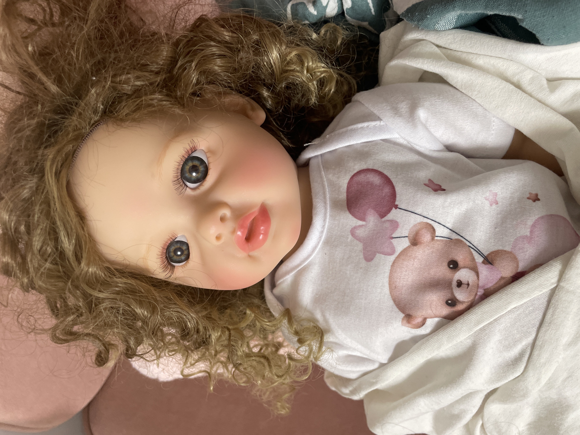 A doll is depicted lying on a soft, light pink upholstered surface, likely a sofa or bed. The doll's head is positioned towards the right side of the frame, with its body extending to the left. It has pale skin, prominent blue-grey eyes with defined eyelashes, and rosy cheeks. Its lips are painted pink and are slightly parted. The doll's hair is wavy and light brown or blonde, appearing synthetic, and is spread across the pink surface. It is dressed in a white long-sleeved garment, possibly a onesie, which features a light pink cartoon bear holding a pink balloon, accompanied by small pink stars. A section of white fabric, likely part of its clothing or a blanket, covers a portion of its upper torso. A small area of patterned green and teal fabric is visible at the bottom left, underneath the doll's clothing. No specific visual cues within the image allow for the identification of the location as the city of Izhevsk, Russia.