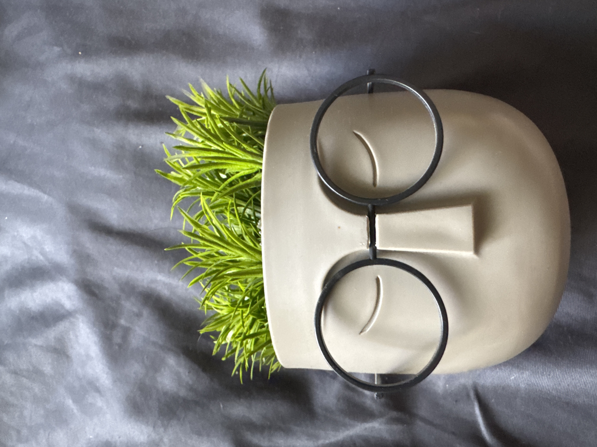 The image shows a close-up, top-down view of a planter shaped like a face with closed eyes and glasses. The planter is light beige and contains bright green artificial grass. The planter is placed on a dark grey, wrinkled fabric surface. The image does not contain any information about the city of Sofia, Bulgaria, or any other specific location.