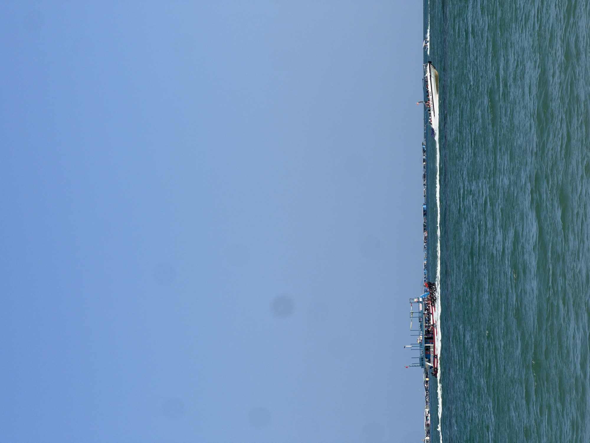This image, captured in Nam Nha Trang, Vietnam, presents a wide vista of a clear blue sky meeting a vast expanse of water, punctuated by a dense line of boats along the horizon. The upper two-thirds of the frame are filled with a pristine, cloudless blue sky, indicating bright, sunny daylight. The lower portion displays dark blue-green water, showing gentle ripples and waves across its surface.
Along the relatively low horizon line, a long procession or gathering of numerous boats of varying sizes and types extends across the entire width of the image. These vessels appear to be either stationary or moving slowly in a coordinated line.
One notable boat, a larger white vessel visible towards the right, has multiple decks and structures, with several people clearly visible on board. The name "MINH HIEU 499" is distinctly legible on its hull, and what appears to be an American flag flies from its stern. Other boats in the line range from smaller, more rustic-looking vessels to other multi-deck boats, suggesting a mix of fishing boats, tour boats, or local transport.
The scene conveys a sense of active marine life or a significant event on the water, possibly a regatta, a tourist excursion, or a daily gathering of the local fleet in Nam Nha Trang. The good weather conditions, clear visibility, and the sheer number of boats imply a lively atmosphere.
