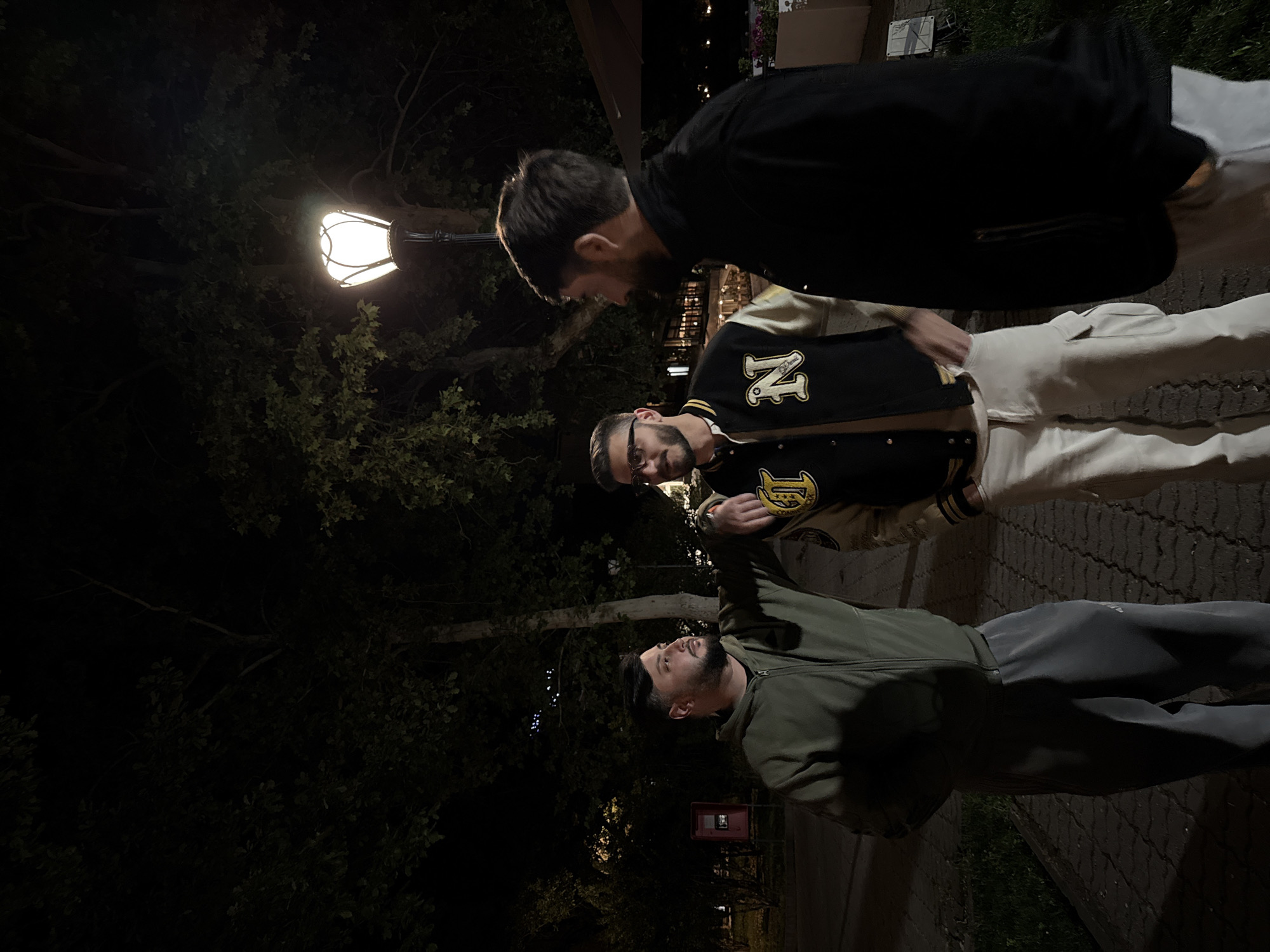 The image shows three men standing on a cobblestone walkway at night. One man is wearing a black jacket and light pants. Another is wearing a varsity jacket with the letter "N" on it, and light pants. The third man is wearing an olive green jacket and dark pants. A street lamp illuminates the scene, with trees visible in the background. A red box is seen near the third man. The location appears to be a park or pedestrian area within a city. Given the known city of Yerevan, Armenia, it's likely that the photo was taken in that location.