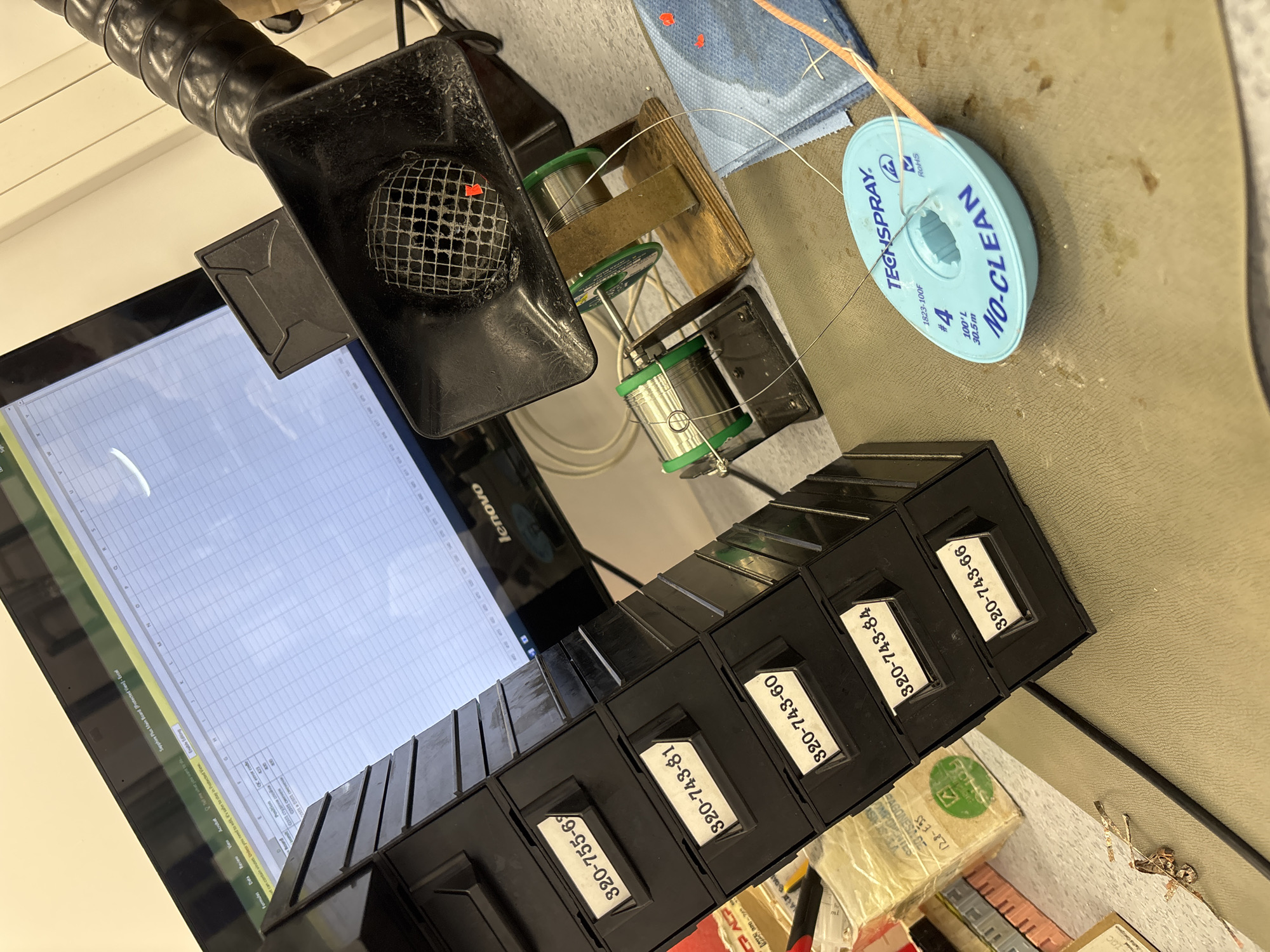 The image depicts a workspace with various electronic components and equipment. In the foreground, a teal-colored spool of solder, labeled "TECHSPRAY NO-CLEAN," is prominently displayed on a textured surface. To the left, a black stack of drawers with labels such as "320-755-69" and "320-743-81" is visible. Behind these drawers, a computer monitor displays a spreadsheet, likely from Microsoft Excel, with rows and columns of data. A Lenovo logo is visible on the monitor's bezel.
In the mid-ground, there is a black fume extractor with a mesh screen, positioned above two spools of solder on wooden stands. The fume extractor has a red mark on its mesh. A thin wire extends from the solder spools and the fume extractor. There are also some small, reddish-orange specks on a blue cloth or paper towel to the right of the solder spools.
In the background, the office environment is suggested by the presence of the computer monitor and what appears to be office supplies or components stacked to the left of the drawers. The lighting suggests an indoor setting, possibly an office or workshop. There are no people visible in the image. The overall scene suggests an active workspace related to electronics assembly or repair.
The image does not contain any information or visual cues that would allow for the determination of its location within the city of Stara Zagora, Bulgaria.