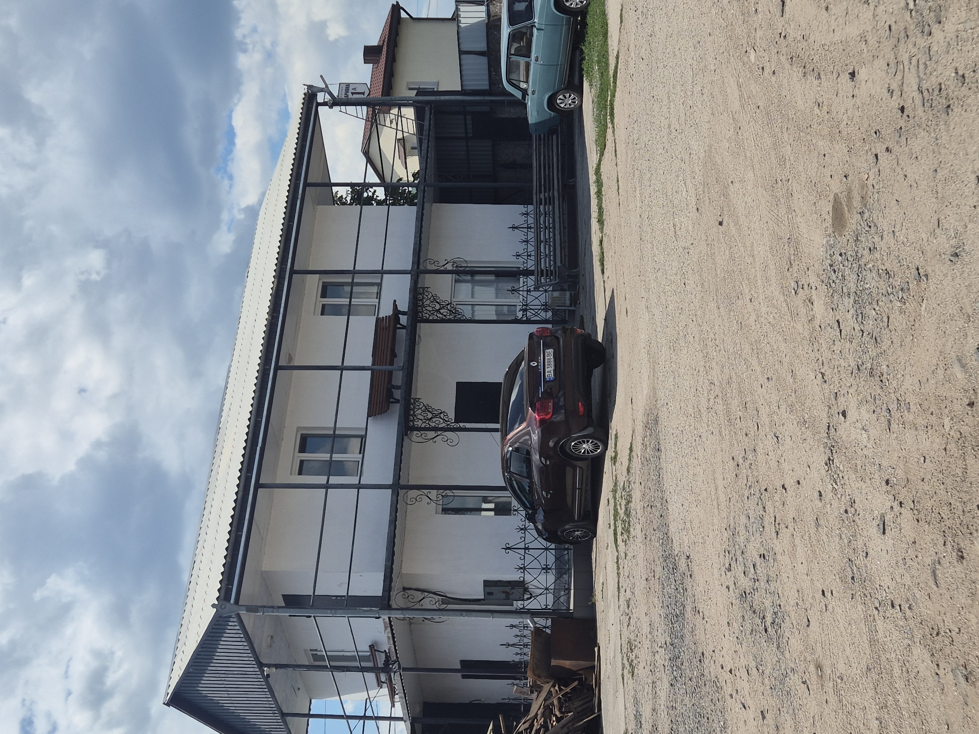 In Pidhaitsi, Ukraine, on a cloudy day, a two-story white building with a corrugated roof stands prominently. A black metal railing encircles the upper story, accessed by an external staircase. Two cars are parked in front of the building on a gravel lot. A dark brown sedan is closer to the viewer, while a light blue older model sedan is parked further away. Piles of wood and other debris are visible to the left of the building. A street sign with Cyrillic lettering, "ВУЛИЦЯ ДАРКОВА 1А" (Dneukova Street 1A), is affixed to the side of the building. The overall scene suggests a commercial or residential property in an outdoor setting.