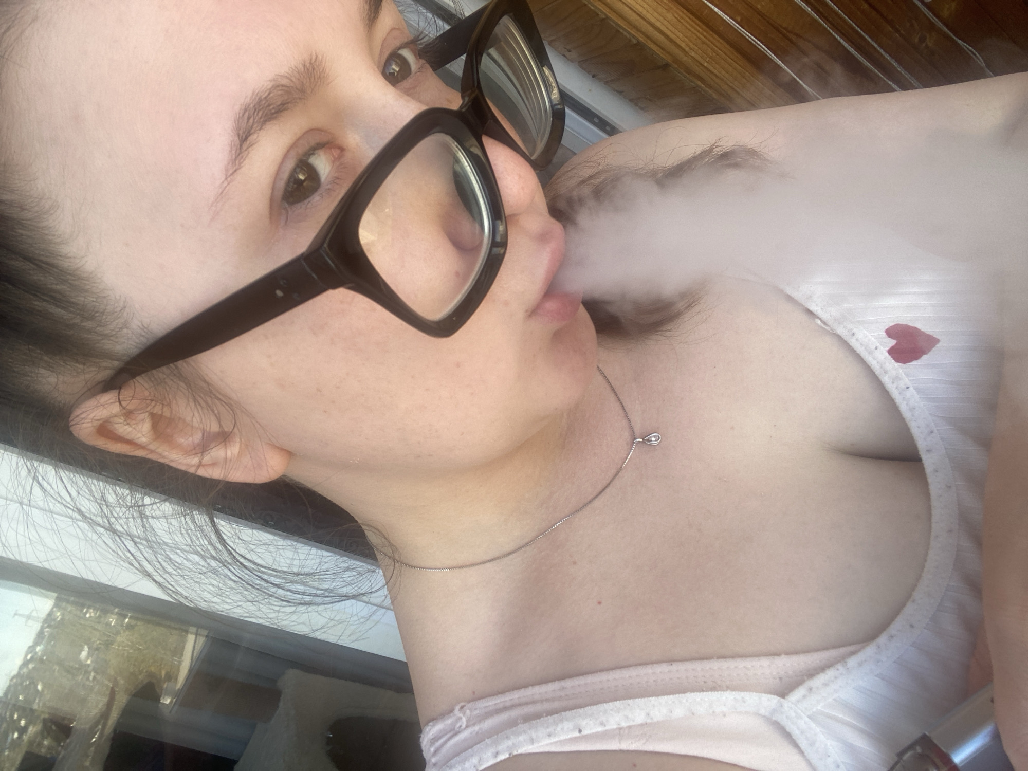 A woman with black-framed glasses is exhaling a large cloud of vapor. She is wearing a white tank top with a small red heart detail and a delicate silver necklace. The scene is indoors, near a window reflecting an exterior view of a building. There is no information in the photo to suggest that this image was taken in the city of Chaikovsky, Russia.
