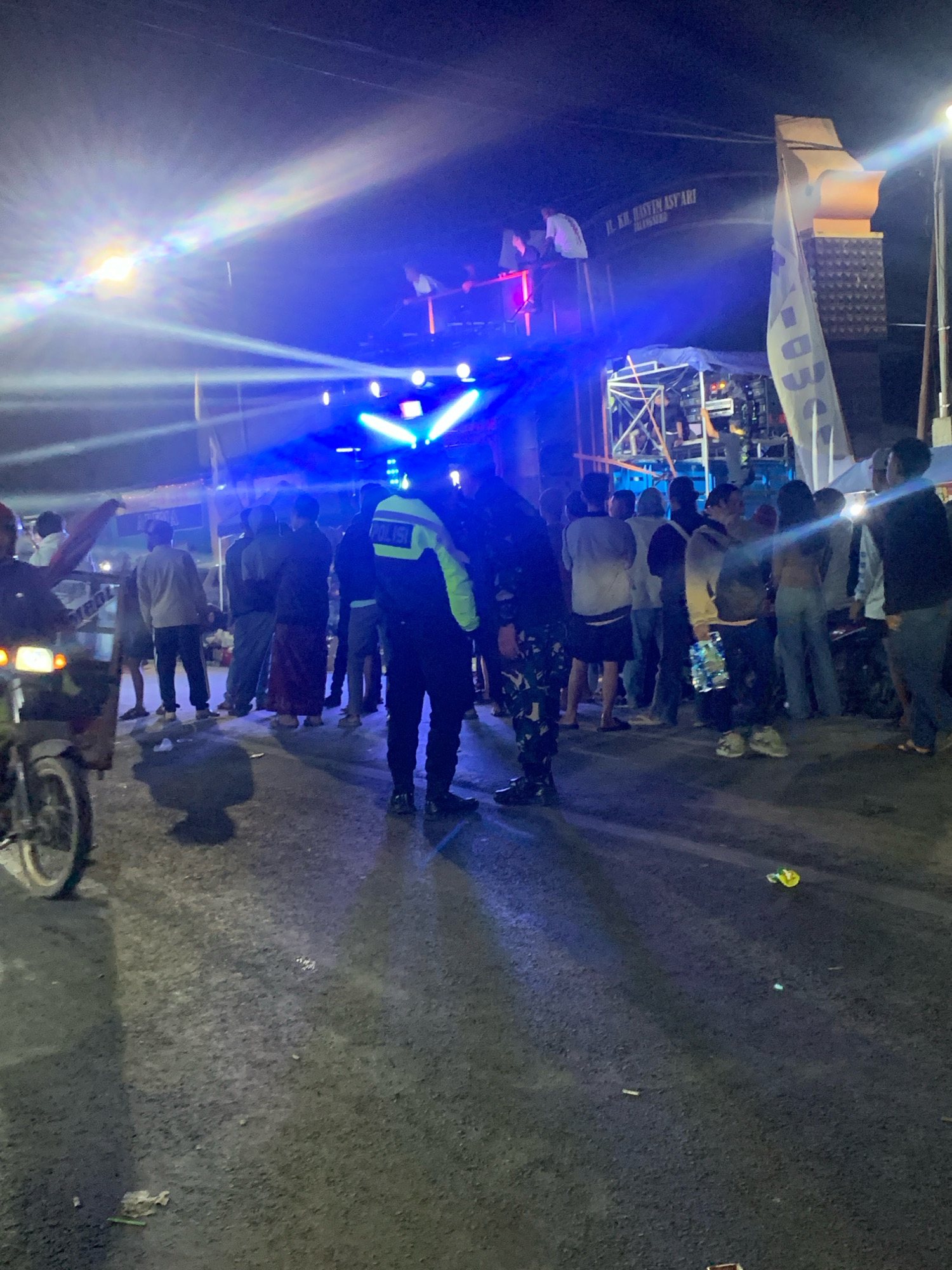 This image depicts a nighttime outdoor scene, likely a street in Talangsuko, Indonesia, illuminated by bright stage lights and possibly vehicle headlights. A crowd of people, numbering at least twenty, are gathered, appearing to be attending an event such as a concert or festival. The lighting creates strong beams and lens flares, casting shadows on the asphalt ground. In the foreground, a motorcycle is partially visible on the left. Several individuals are wearing casual clothing, while one person in the center is wearing a dark uniform with "POLISI" visible on the back, indicating a law enforcement presence. In the background, a stage or setup with speakers and lighting equipment is prominent, with some people standing on or near it. A banner with text, partially obscured, is also visible. The overall atmosphere suggests a lively public gathering.