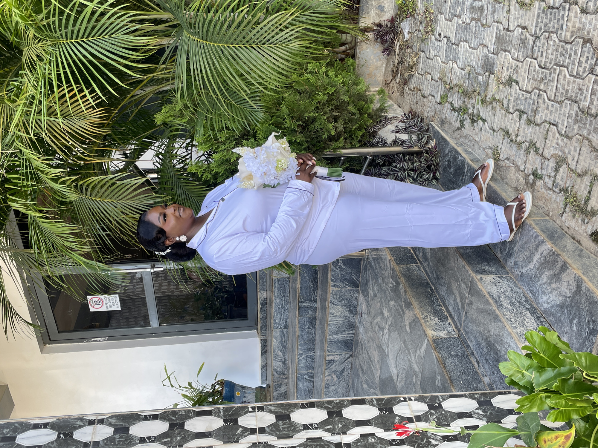 A woman in white pajamas and flip-flops stands on gray stone steps outside a building, holding a bouquet of white flowers. She is surrounded by lush green foliage, including a large palm tree. The building has a gray and white exterior with a glass door or window displaying a sign. The ground is covered in interlocking gray pavers. The location is Jos, Nigeria, evidenced by the environment and likely the individual's ethnicity.