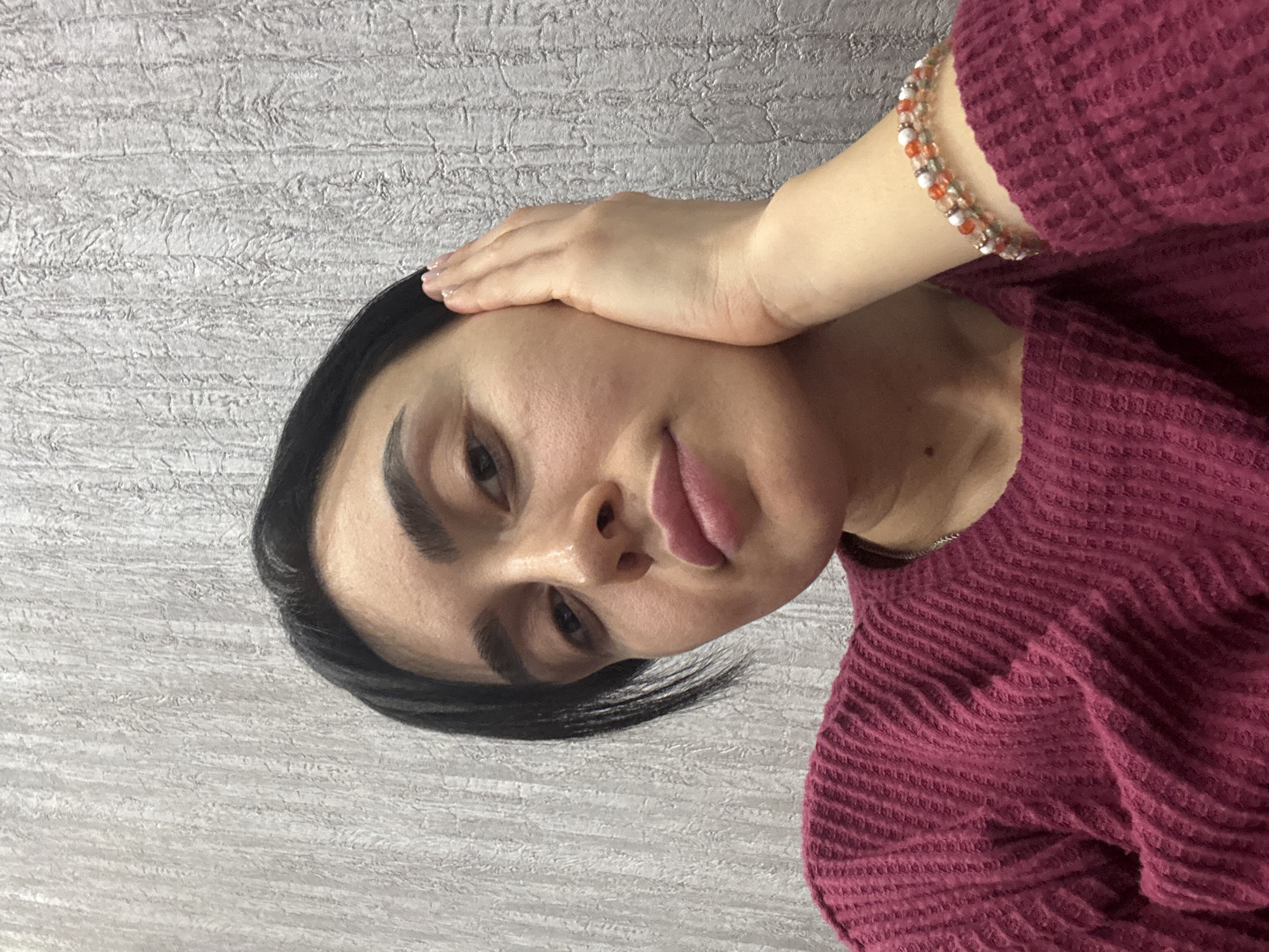 The image displays a close-up portrait of an adult female, positioned from the chest up, slightly off-center to the left. She has short, dark hair, dark eyebrows, and dark eyes, looking directly at the camera. Her right hand is raised, with fingers gently pressed against her right cheek and ear area. She is wearing a burgundy, waffle-knit, long-sleeved sweater. A thin, silver-colored chain is visible around her neck, and a multi-strand beaded bracelet featuring clear, orange, white, and green beads is on her right wrist. The background consists of a light grey wall with a textured, irregular vertical pattern. No visual information within the image indicates a specific location in Tbilisi, Georgia.