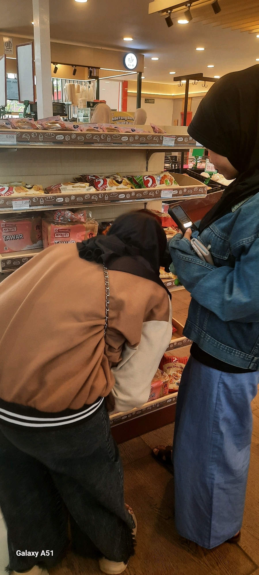 Two individuals, both wearing hijabs, are browsing the shelves of a bakery or convenience store. The person on the left is bending down, looking at items on a lower shelf, and is wearing a brown bomber jacket over dark pants. A silver chain strap is visible on their jacket. The person on the right is standing and looking at their mobile phone, holding a wallet in their left hand. They are wearing a blue denim jacket over a light blue skirt or pants. The shelves are stocked with various packaged bread and pastry products. The store environment appears well-lit with overhead lighting and has a clean, tiled floor. The store is located in the city of Wlingi, Indonesia.