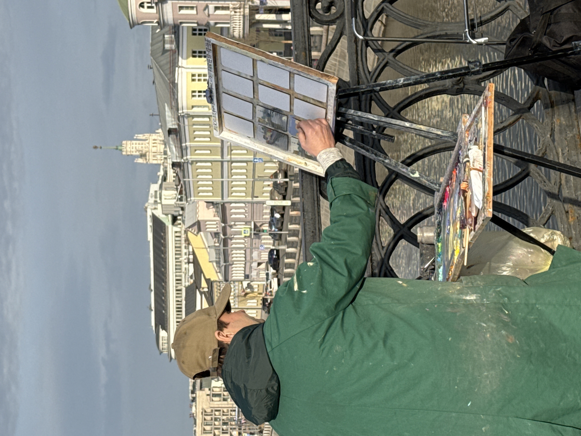 An individual, viewed from behind, is painting on a bridge over the Moskva River in Moscow, Russia. The person, appearing male, wears a brown baseball cap and a dark green, paint-stained long coat. They hold a paintbrush to one of twelve small rectangular canvases framed within a larger wooden structure on a black metal easel. Below the canvas, a wooden palette displaying various oil paints and tubes is attached to the easel. The bridge features an ornate, dark metal railing with circular patterns. Across the water, an embankment road with parked and moving vehicles fronts a series of multi-story buildings, predominantly in pastel yellow and pink hues, with numerous windows. In the distant background, the tall, light-colored spire of the Kotelnicheskaya Embankment Building is visible against an overcast sky.