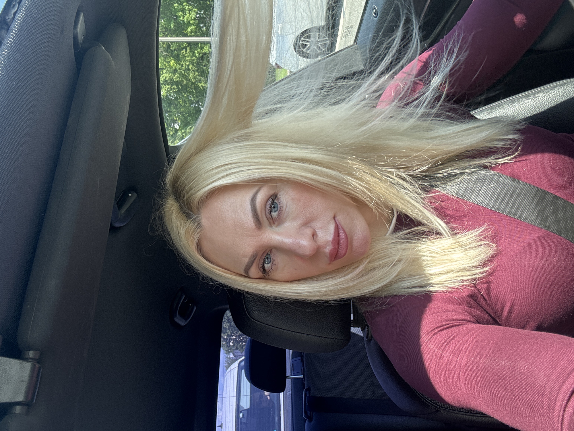 A woman with blonde hair is sitting in the driver's seat of a car. She is wearing a maroon long-sleeved shirt and a seatbelt. Her hair is being blown by the wind from an open window. She is looking at the camera with a slight smile. The car's interior is visible, including the dashboard and part of the passenger seat. Outside the car, trees and other vehicles can be seen. The scene appears to be in Saint Petersburg, Russia, given the context.