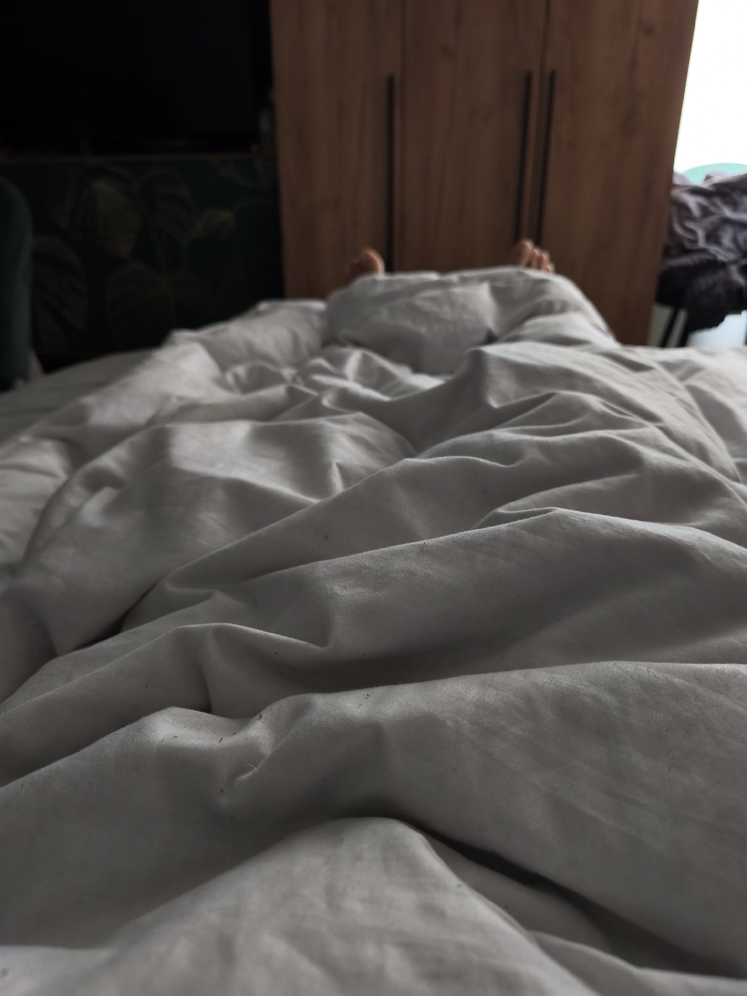 A close-up shot presents a rumpled gray bedsheet covering a person. Two feet are slightly visible at the head of the bed, suggesting someone is lying underneath. A wooden wardrobe with dark handles stands blurred in the background. To the left, a dark green object with a patterned surface is partially visible, along with a darker, crumpled item to the right. The location is not discernible from the image.