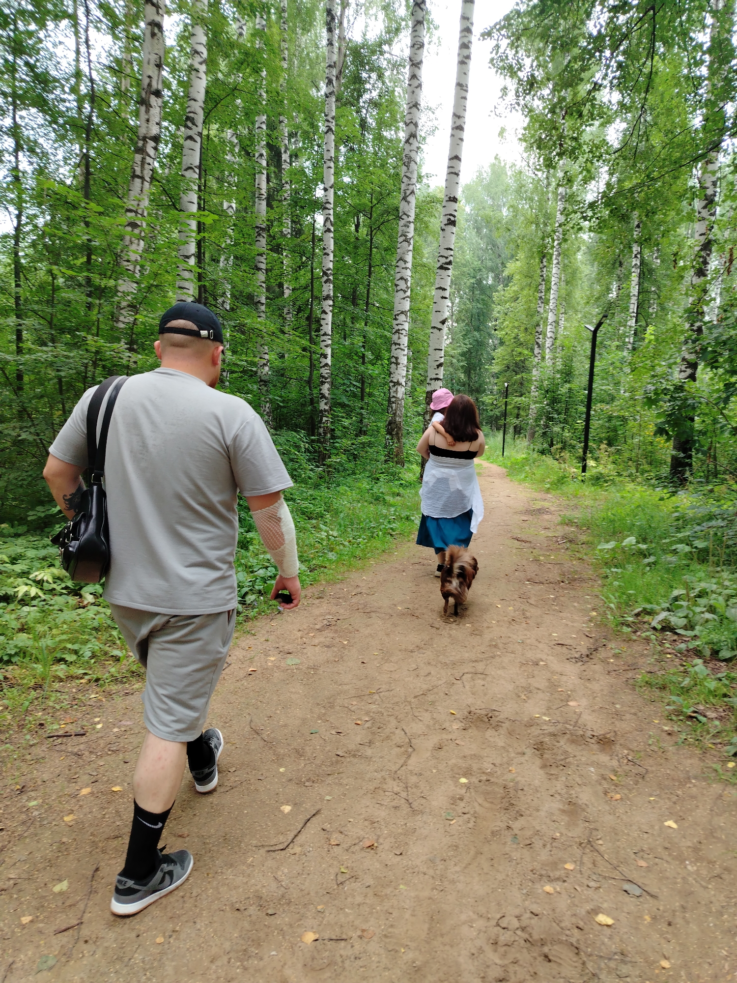 A man wearing a grey t-shirt, grey shorts, black socks, and grey sneakers walks on a dirt path in a birch forest in Noviy, Russia. He has a black baseball cap on and a black bag slung over his right shoulder. His left arm is bandaged. A woman wearing a blue skirt and a white top walks ahead of him, carrying a child on her hip. A small dog walks in front of the woman. The path is lined with birch trees and green foliage.