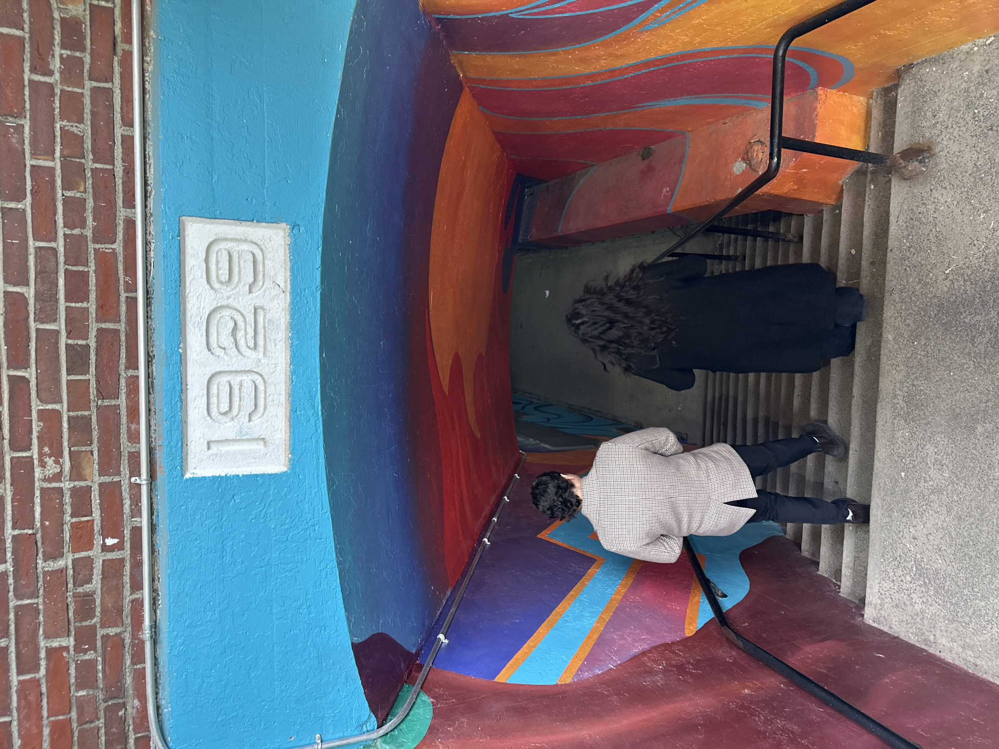 This media file captures a vibrant, artistically decorated staircase leading into a tunnel or underpass, located in the Village of Cold Spring, United States of America. The walls and ceiling of the passage are adorned with a colorful mural featuring abstract, wavy patterns in shades of blue, purple, red, orange, and yellow.
On the left side of the entrance, a section of a red brick wall is visible, adjacent to a brightly painted light blue wall. A white rectangular plaque with the embossed number "629" is affixed to this blue wall. A silver metal conduit runs along the brickwork and then horizontally along the lower portion of the blue wall, extending into the tunnel.
Two individuals are present on the concrete staircase. The person further down the stairs is seen from behind, wearing a dark coat and having dark, curly hair. The second person, closer to the entrance, is also viewed from behind, wearing a light plaid jacket and dark pants, seemingly looking down the stairs as if preparing to descend. A black metal handrail guides the descent on the right side of the staircase. The lighting suggests it is daytime, with natural light illuminating the entrance of the passage.