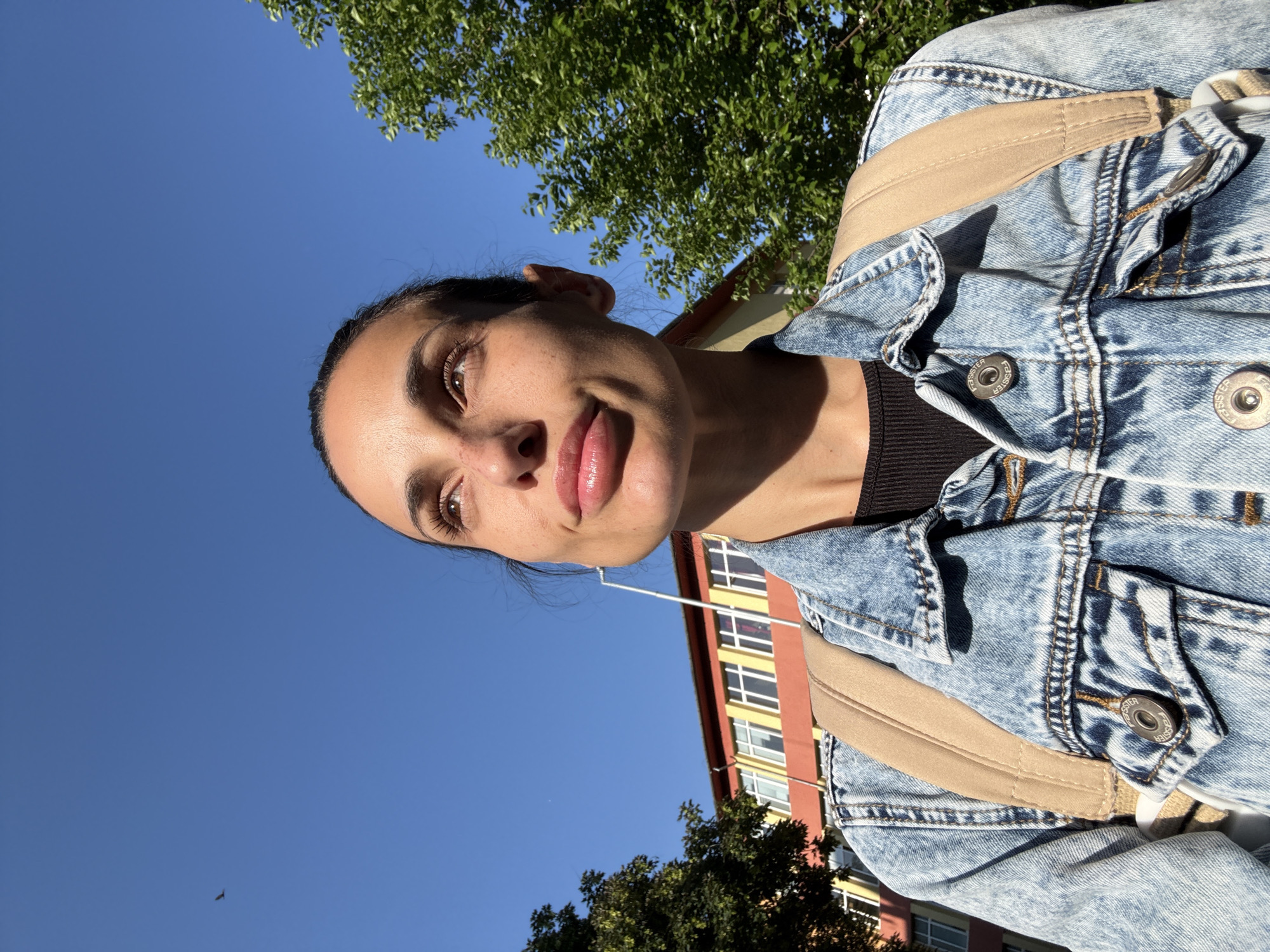 A selfie-style photograph of a young woman with olive skin, brown eyes, and dark hair pulled back, wearing a blue denim jacket over a black top and a tan backpack. She is smiling slightly. The background includes a clear blue sky, green trees, and a red and yellow building. Based on the architectural style and color scheme of the building, the location is likely in Pleven, Bulgaria.