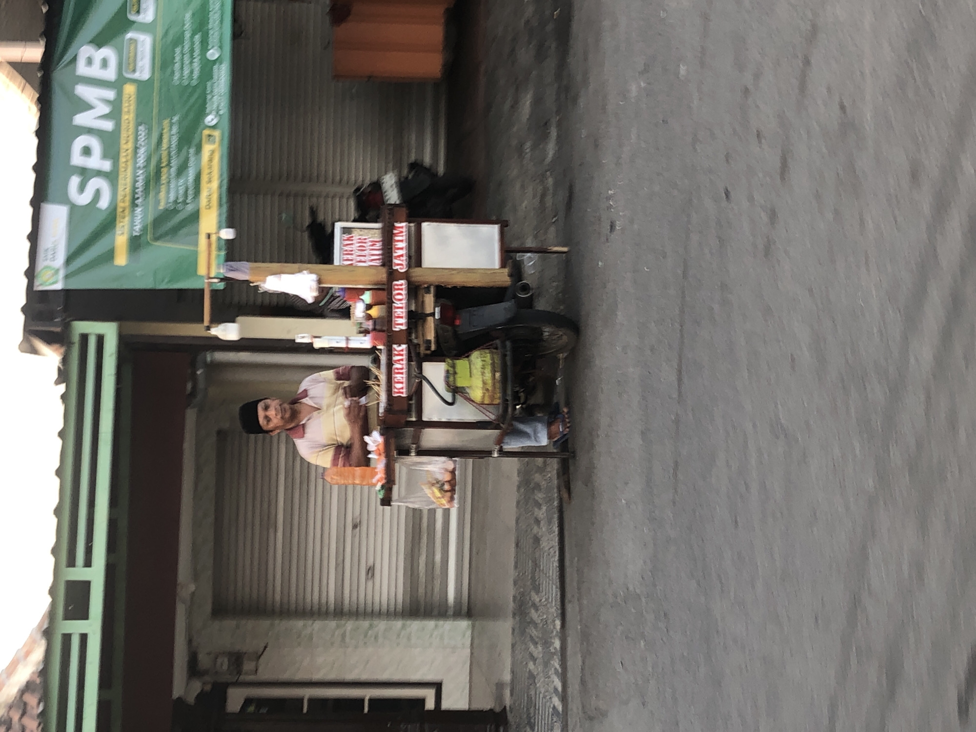 A street vendor stands behind his cart, which is attached to a motorcycle, in front of a closed storefront. The vendor, a man wearing a black cap and a striped shirt, is holding something in his hands. The cart has various items on it, including bottles of sauce, raw eggs in a plastic bag, and skewers of food. A gas cylinder is visible on the side of the cart. In the background, a large green banner with the text "SPMB" is displayed, along with other writing in Indonesian. The storefront behind the vendor has a rolled-down metal door. The scene appears to be on a street in Glagah Arum, Indonesia.