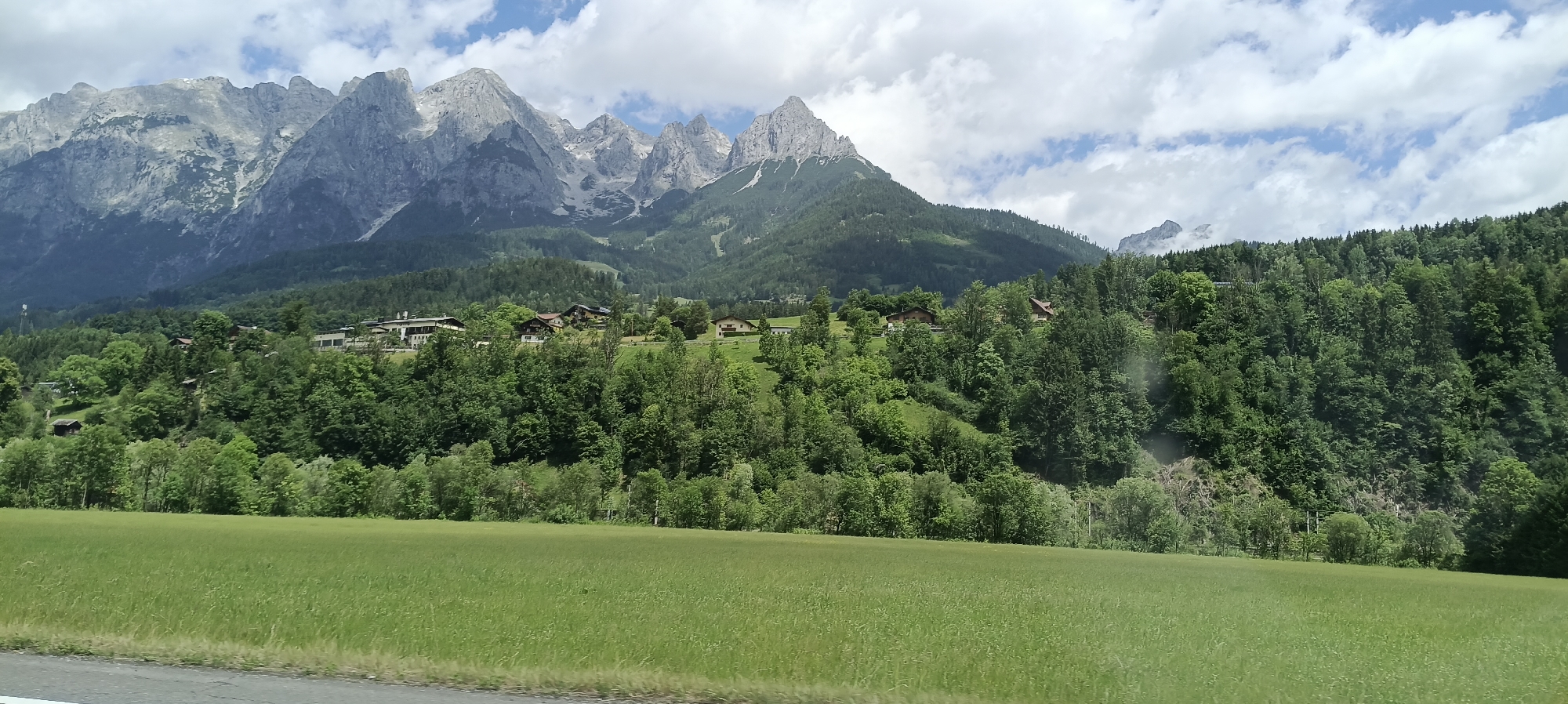 The image captures a scenic view of the Bischofshofen area in Austria. In the foreground, a lush green field stretches across the frame. Behind the field, a dense forest blankets the lower slopes of the mountains. Scattered throughout the forest are several traditional Austrian houses, their roofs a mix of dark wood and lighter tiles. The background is dominated by majestic, rugged mountains with grey, rocky peaks, some of which are capped with snow. The sky above is partly cloudy, with white clouds drifting across a pale blue expanse. The overall impression is one of a serene alpine landscape with human habitation integrated into the natural beauty.