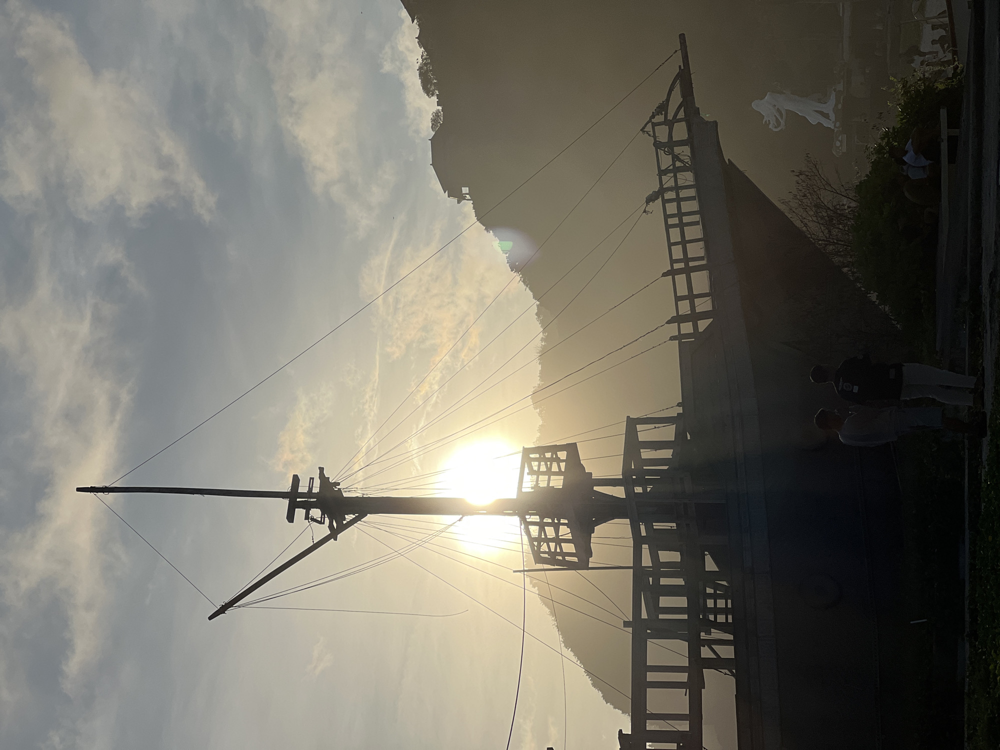 The image shows a silhouette of a large ship, likely a replica, with a tall mast and rigging against a bright sun. The ship is located in Kuta Selatan, Indonesia. The sun is partially obscured by the ship's structure and a mountain in the background. Two individuals are standing on the right side of the ship, near some greenery. Above the ship, there is an indistinct white sculpture against the mountain. The sky is partly cloudy.