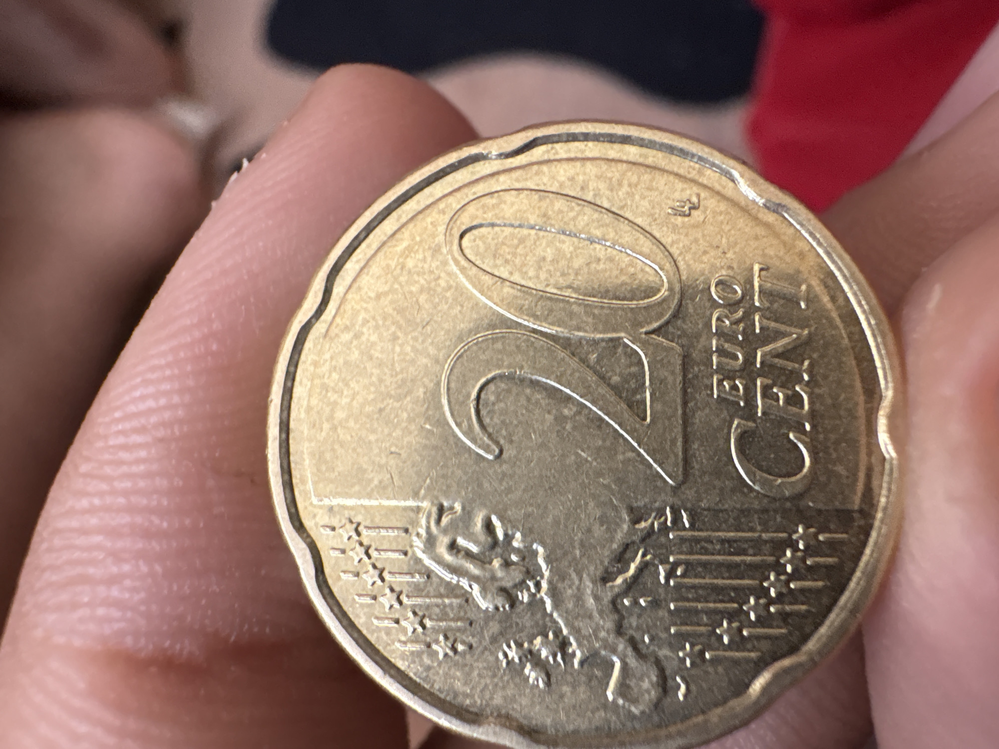 A hand holds a 20 Euro cent coin. The coin shows the numeral "20" and the words "EURO CENT". Below the numeral and text is an image of Europe. The location is unknown.