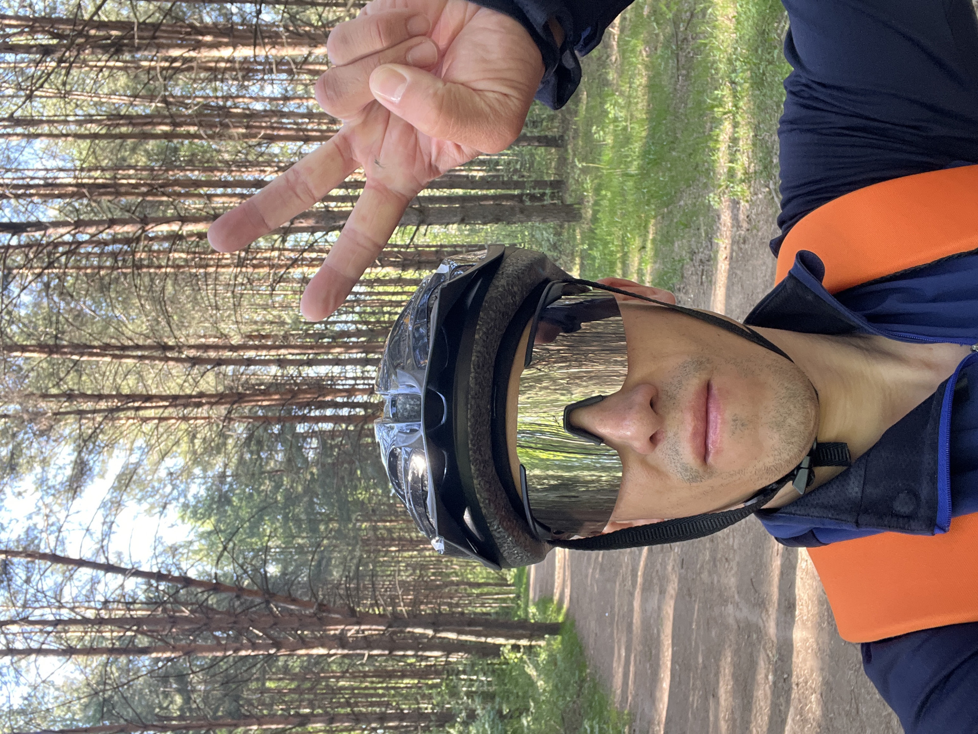A man wearing a black helmet and mirrored sunglasses poses for a selfie in a forest, holding up a peace sign with his left hand. He's wearing a navy blue jacket with an orange band underneath. The background consists of tall pine trees and a dirt path, suggesting a wooded trail. The location appears to be in a natural area, possibly within or near the city of Chaikovsky, Russia, given the prevalence of coniferous forests in that region.