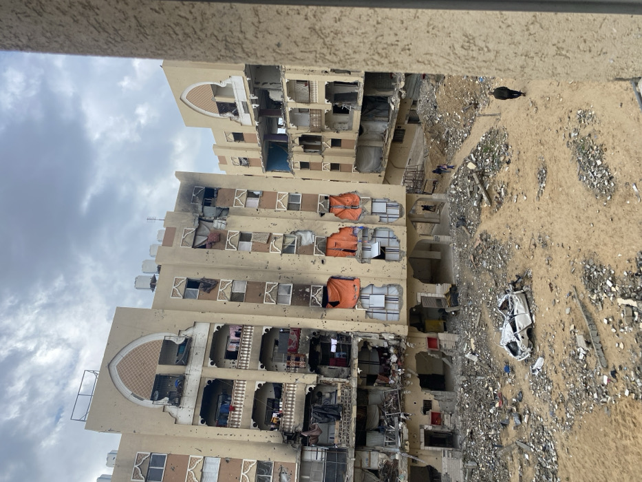 This image depicts a scene of widespread destruction in Khan Yunis, Palestinian Territory, captured from an elevated perspective. The foreground features a vast expanse of debris-strewn sandy ground, encompassing rubble, broken concrete, and discarded materials. A heavily damaged, white four-door vehicle, appearing crushed and stripped, lies prominently in the lower-left quadrant of this area.
Three multi-story residential buildings, constructed with light brown facades and featuring distinctive arched decorative elements on upper floors, dominate the middle ground. All structures exhibit extensive damage:
* **The leftmost and central buildings** show numerous projectile holes peppered across their facades. Many windows are shattered or missing, exposing darkened interiors. Sections of walls are blown out, revealing mangled rebar and collapsed floors. Burn marks, indicative of fire damage, are visible on the upper stories of the leftmost building. Orange tarpaulins cover multiple damaged sections on a lower floor of the central building. Balconies are either destroyed or heavily damaged, with remnants of clothing or fabrics hanging from some. Water tanks are visible on the roofs of these buildings.
* **The building on the right**, partially obscured by the structure from which the photo was taken, also displays significant structural damage, with entire sections of walls missing and interiors exposed.
Several individuals are present on the ground amidst the debris. One person, dressed in dark clothing, walks from right to left on a sandy path in the lower-right foreground. Further back, near the base of the central building, two smaller figures are visible walking on a similar path, with another distant figure discernible behind them. The sky above is overcast with a mixture of grey and white clouds. A vertical brown structure, likely a window frame or pillar of the vantage point building, partially frames the scene on the right.
