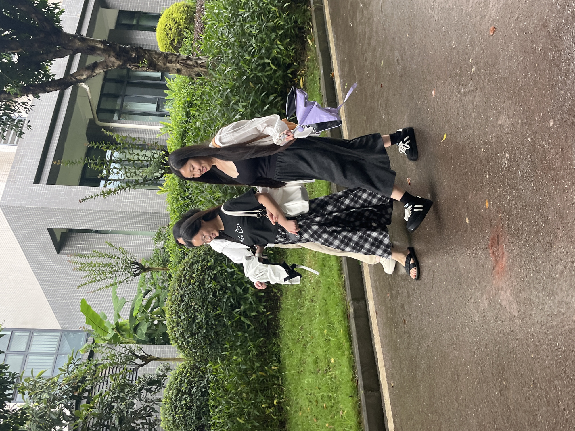 Three young women are walking on a wet paved surface in front of a building in Jiangjin, China. The woman in the foreground is wearing a black tank top, a white open-front shirt, a long black skirt, and black sneakers. She is holding a folded purple umbrella. To her left, a woman in a black t-shirt and a black and white plaid skirt is walking. She is carrying a white tote bag. Another woman, mostly obscured, is walking behind them. The building behind them is multi-story with modern architecture and large windows. Green foliage, including bushes and trees, surrounds the building and the walking path. The ground is wet, suggesting it has recently rained, with some scattered leaves visible. The overall scene depicts a casual walk on a university campus or similar urban environment.