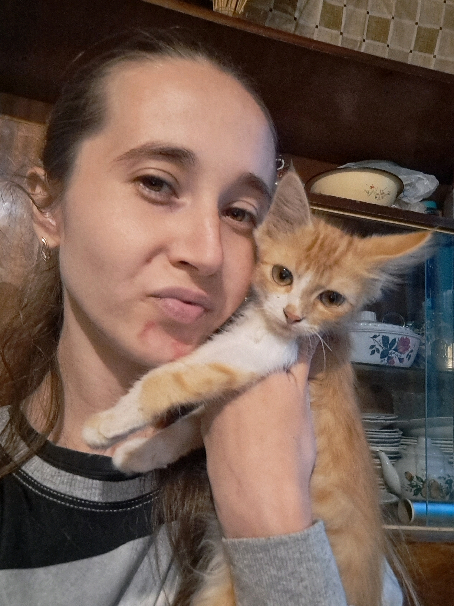A young woman with brown hair pulled back and wearing a grey and black striped shirt holds an orange and white kitten. The kitten is small with large ears and amber eyes. The woman's skin appears to have some redness on her chin. In the background, a wooden cabinet or shelf unit is visible, containing various dishes and decorative items. The image is a selfie, taken indoors, with the woman and kitten as the primary subjects. The scene is likely within a residential setting in the city of Vypasne, Ukraine.