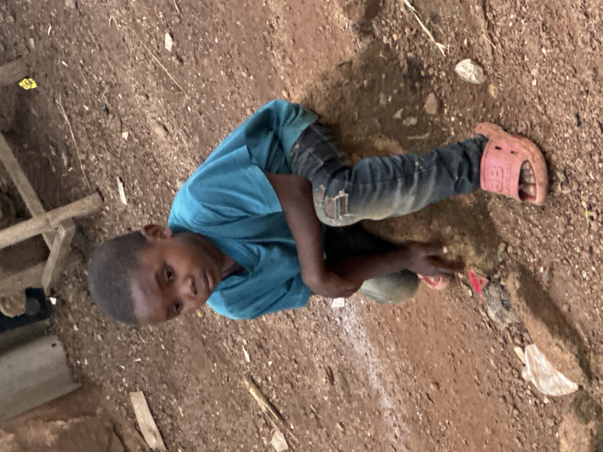 In Gura Top, Nigeria, a young child sits on a rock amidst a dirt ground. The child is wearing a teal t-shirt and ripped blue jeans, complemented by pink slides. The background consists of dirt, rocks, and some debris, with a partially visible wooden structure. The overall scene depicts a simple, outdoor environment.