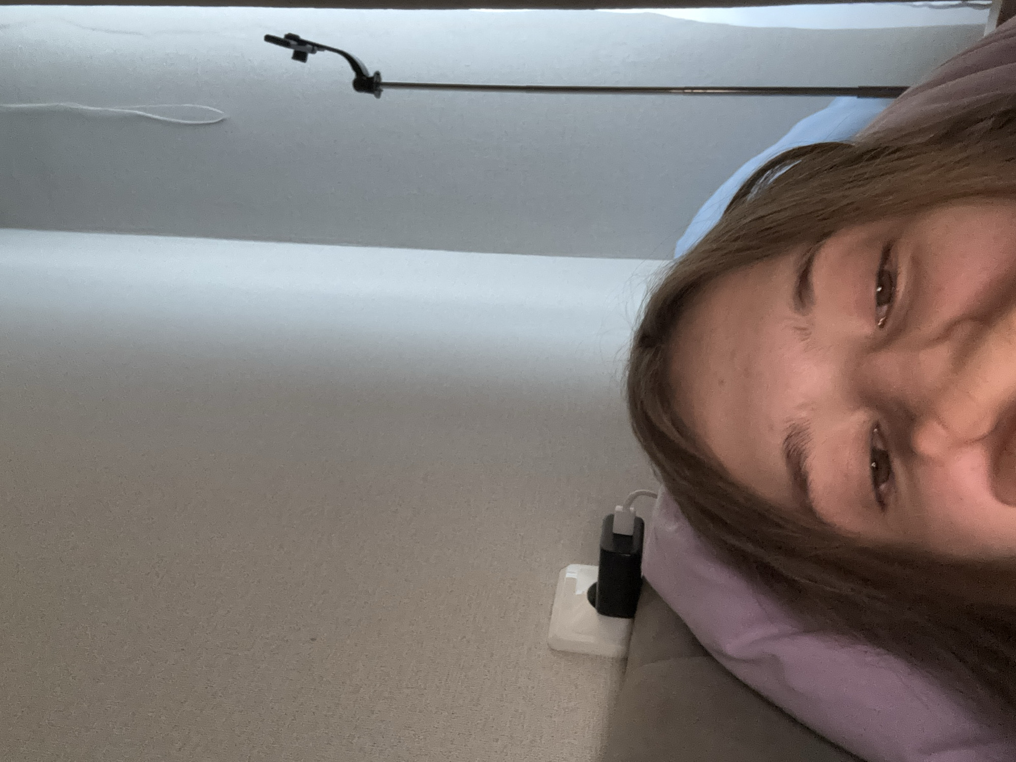 The image shows a close-up, upside-down selfie of a person lying on a bed. The person's face is the primary subject, with their eyes and forehead visible. They appear to be looking towards the camera. In the background, a light-colored wall can be seen, along with a power outlet with a charger plugged into it. To the right, a tall, thin lamp with a curved arm and a light fixture is positioned against a wall. The environment suggests an indoor setting, possibly a bedroom. The exact location within the city of Unknown, Unknown cannot be determined from the image.
