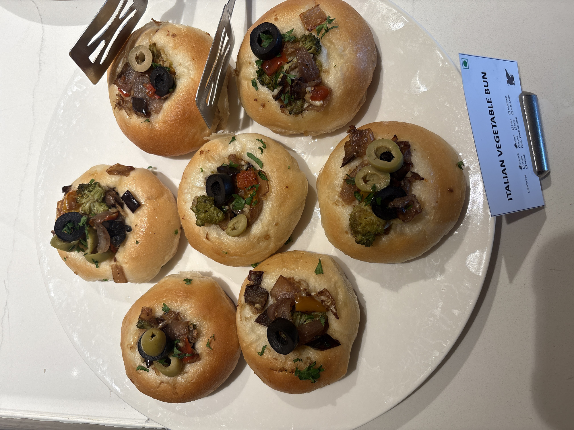 A white, oval platter holds six Italian vegetable buns, each topped with chopped vegetables and olives. The buns are arranged in two rows, with three in the back row and three in the front row. A small, blue and white sign is placed in front of the platter, which reads "ITALIAN VEGETABLE BUN" and lists ingredients. Two silver serving tongs are also visible on the platter. The platter is set on a speckled white countertop. The lighting is bright, creating some glare on the platter.