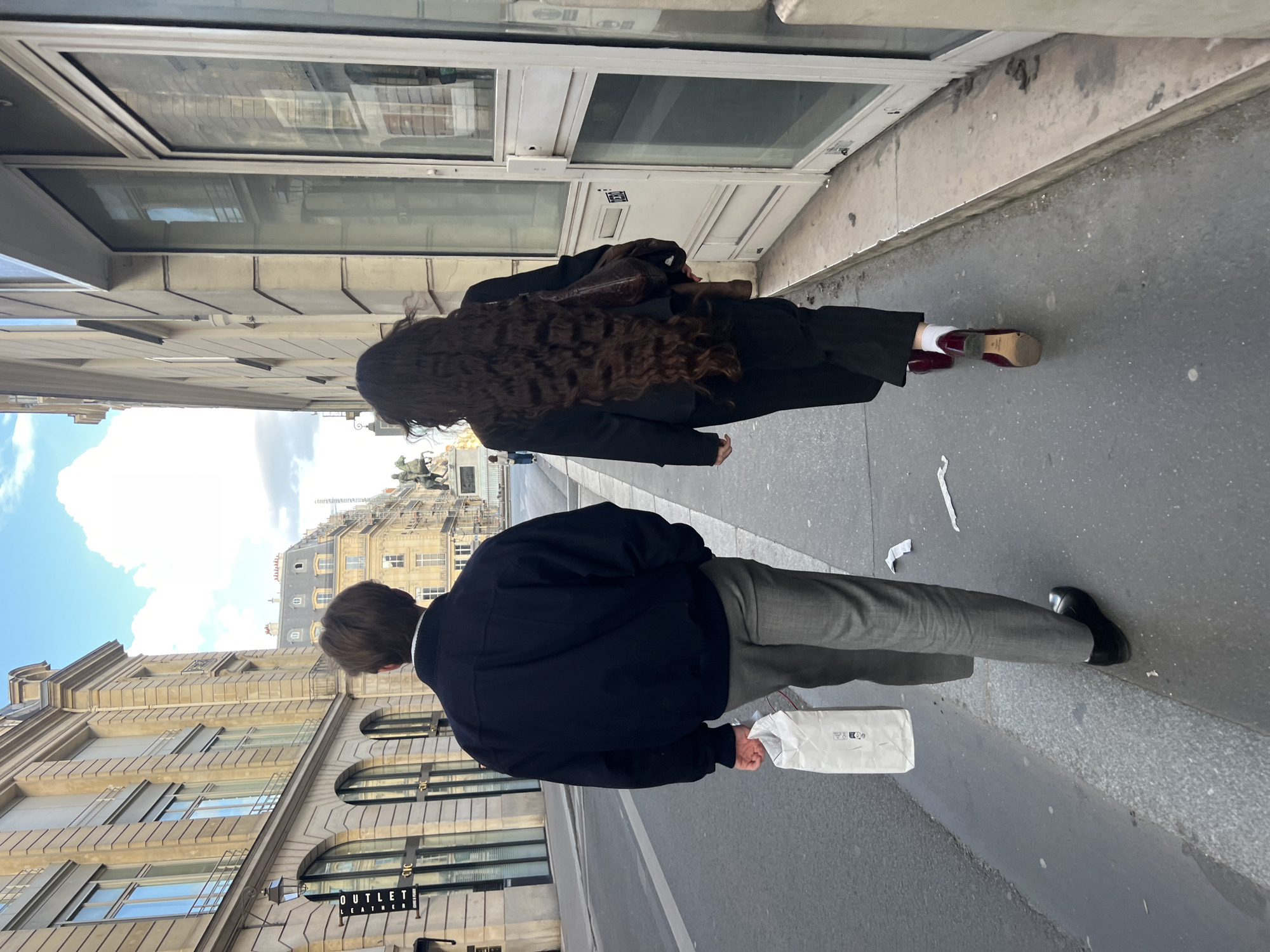 Two people walk down a sidewalk. One person with long, dark curly hair is wearing a black coat, black pants, white socks, and dark red shoes. This person also carries a brown bag. The other person, with short brown hair, wears a dark blue jacket and grey pants. This person is holding a white bag. The sidewalk is next to a building with storefront windows. Across the street, there is a building with a sign that reads "Outlet Leather." The buildings suggest this is Paris, France.