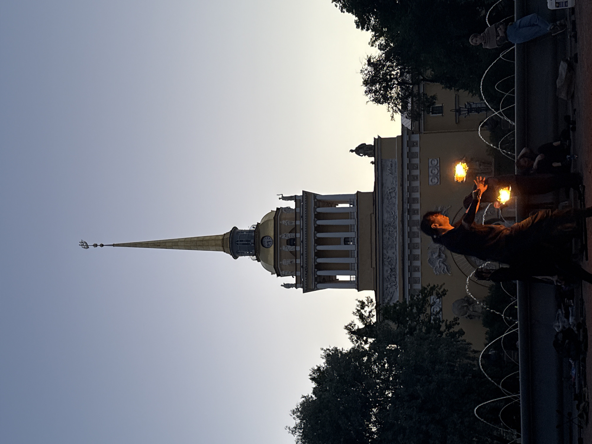 The image depicts a twilight scene in front of the Admiralty Building in Saint Petersburg, Russia. A fire performer is actively engaged in their act, holding flaming props in their hands. To the right, a person sits leaning against a fountain's edge, observing. In the foreground, various discarded items are scattered on the ground. The background features the iconic Admiralty Building with its prominent spire, surrounded by dark trees and illuminated by the fading light of dusk. The fountain in the middle ground displays multiple arcs of water.