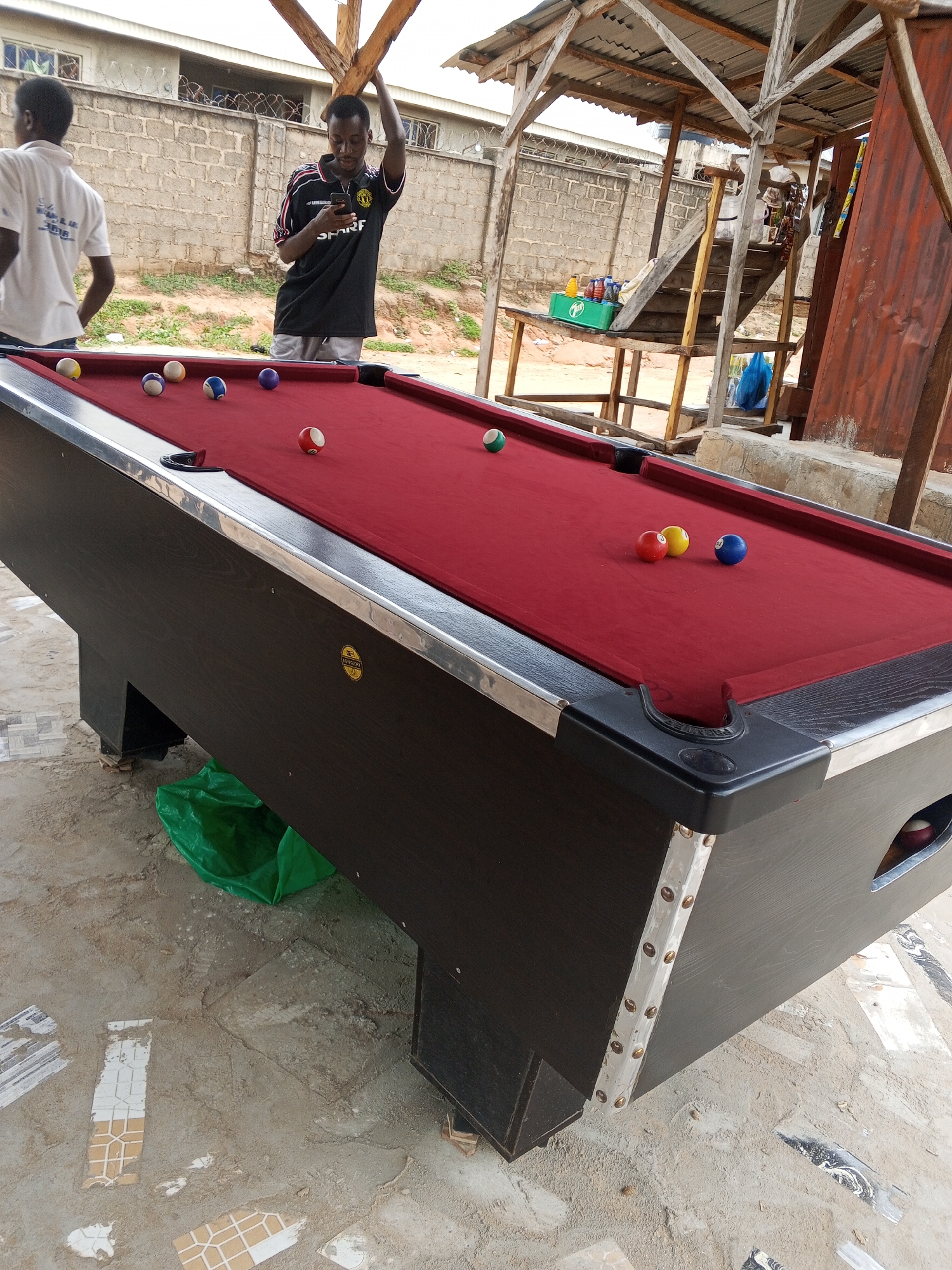 This image captures an outdoor scene at what appears to be a casual gathering spot. In the foreground, a red billiard table with a polished black wooden frame is prominently displayed. Several billiard balls are scattered across the table, including a green ball, a yellow ball, a blue ball, and a red and white striped ball. The table has a metallic trim and visible pockets.
In the background, two young men are present. One man, wearing a white t-shirt, stands with his back to the camera. The other man, dressed in a black and red athletic shirt and grey shorts, is looking at his mobile phone. Behind them, there is a rustic wooden structure with a corrugated metal roof, suggesting an open-air shelter. A set of wooden stairs leads up to a platform within this structure, where various items, including bottles and a green crate, are visible. The ground is made of dirt and scattered tiles, and a green plastic bag lies near the billiard table. A brick wall and barbed wire fence are visible further in the background, hinting at a somewhat urban or semi-urban environment. The overall impression is of a laid-back, perhaps informal, recreational area. The time of day appears to be daytime, with soft lighting. The location cues suggest this is in Lokoja, Nigeria.