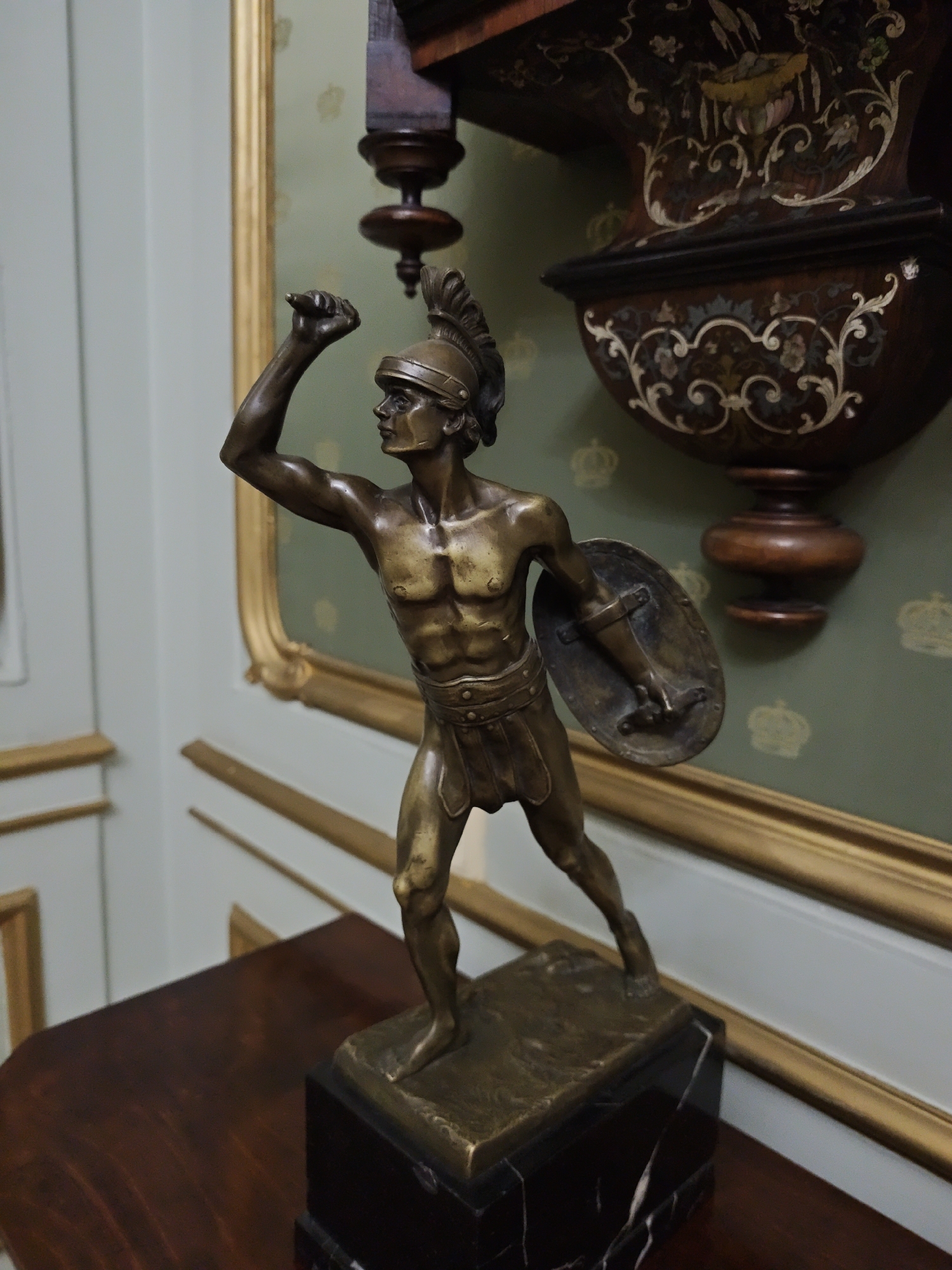 A bronze statue of a Roman soldier stands on a dark marble pedestal, which rests on a polished mahogany surface. The statue depicts a muscular male figure wearing a plumed helmet and a short tunic, holding a shield in his left arm and raising his right arm. The background features a green wallpaper with a repeating crown motif, and a portion of an ornate wooden piece of furniture with inlaid decoration. A golden frame borders the wallpaper. The image appears to be taken indoors, likely in a museum or private collection.